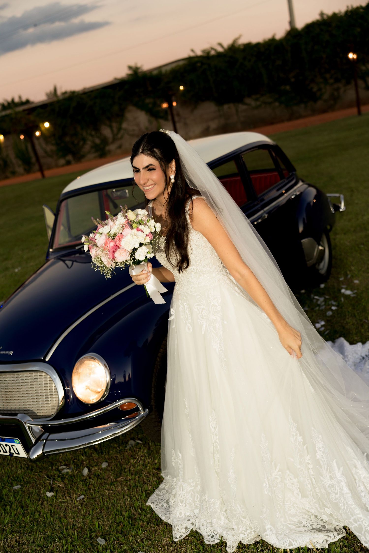 Bride in white lace gown and veil holds pink bouquet, posing beside a vintage navy car at dusk.