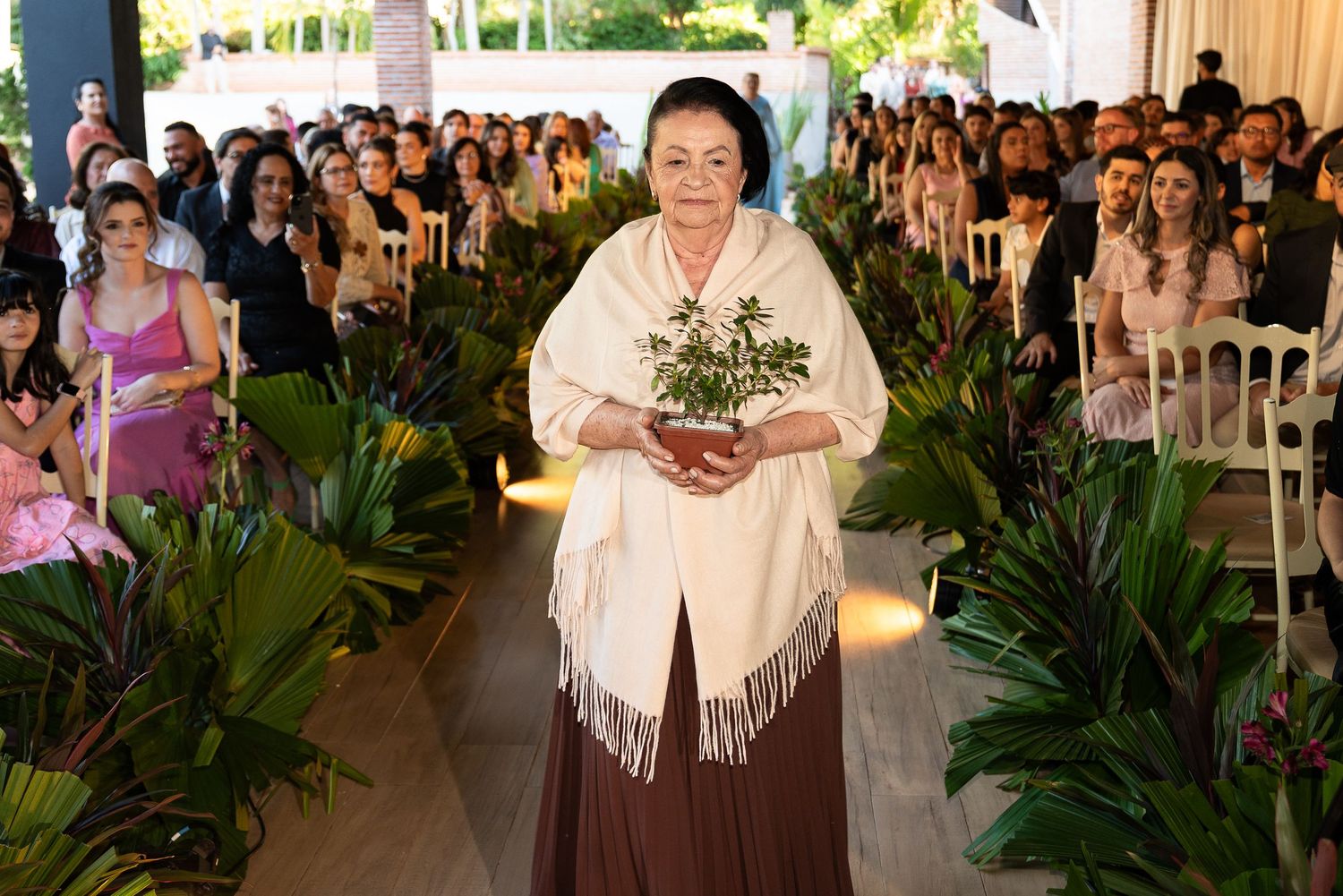 Elderly woman in white shawl carrying green plant walks down aisle at elegant outdoor event with guests seated.