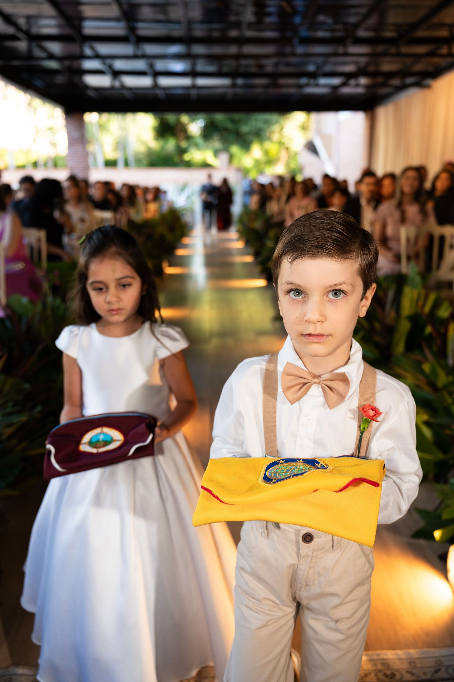 Two children serve as ring bearer and flower girl walking down a wedding aisle, boy in bow tie holding a yellow pillow.