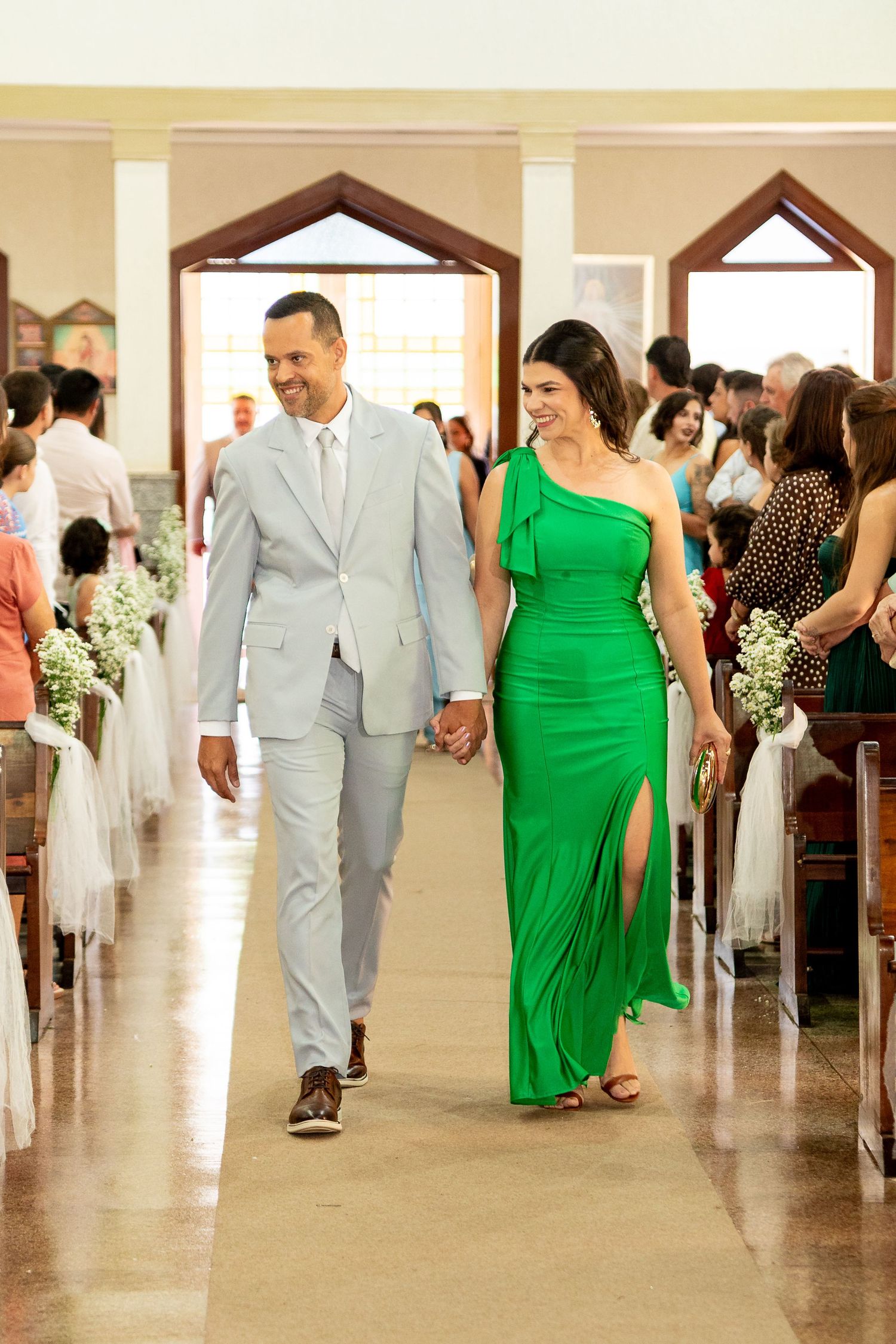 Couple walks down church aisle, man in light gray suit, woman in elegant green one-shoulder gown, wedding ceremony.