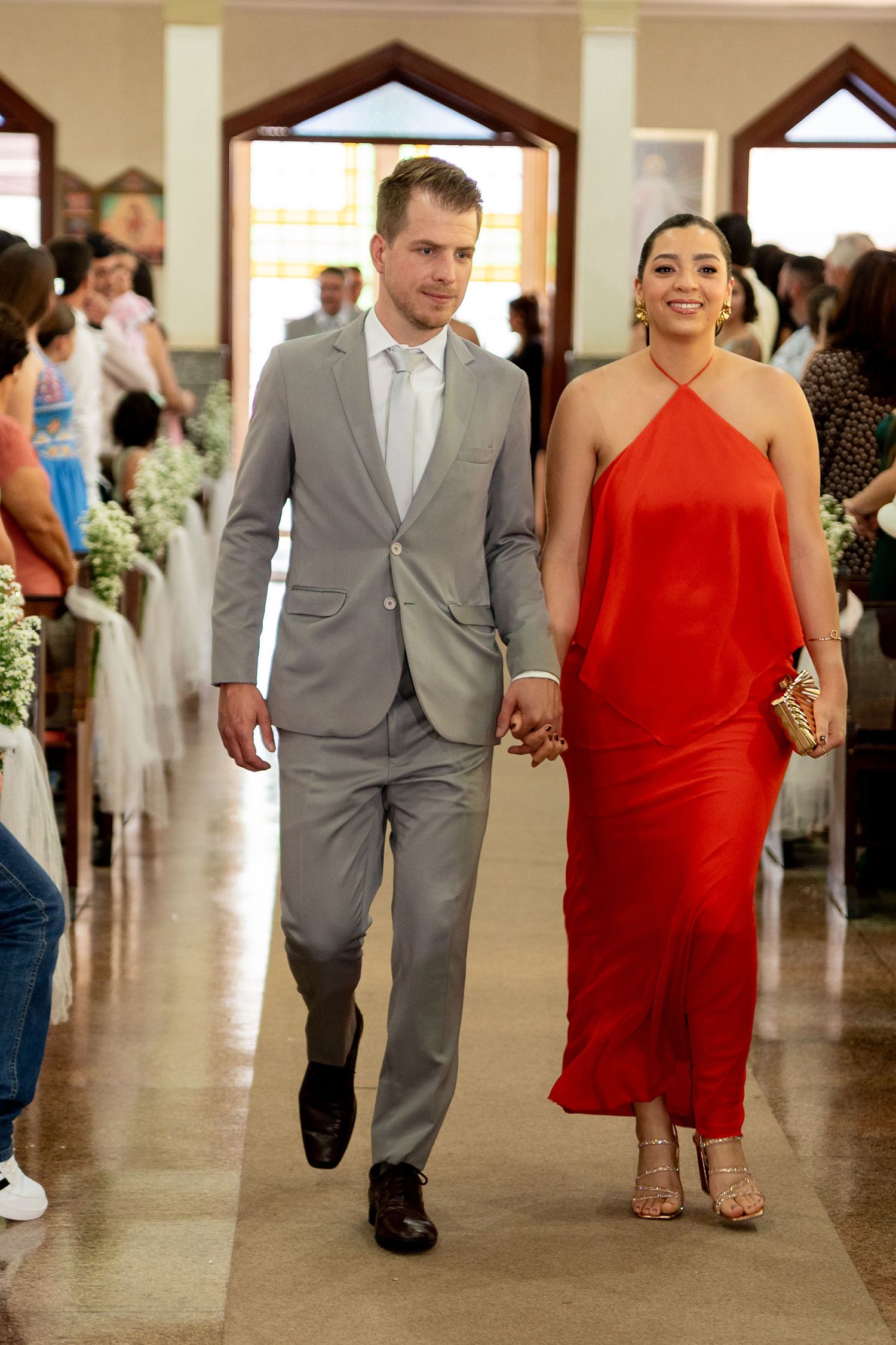 Couple walking down church aisle, man in gray suit, woman in red halter dress, holding hands at wedding ceremony.