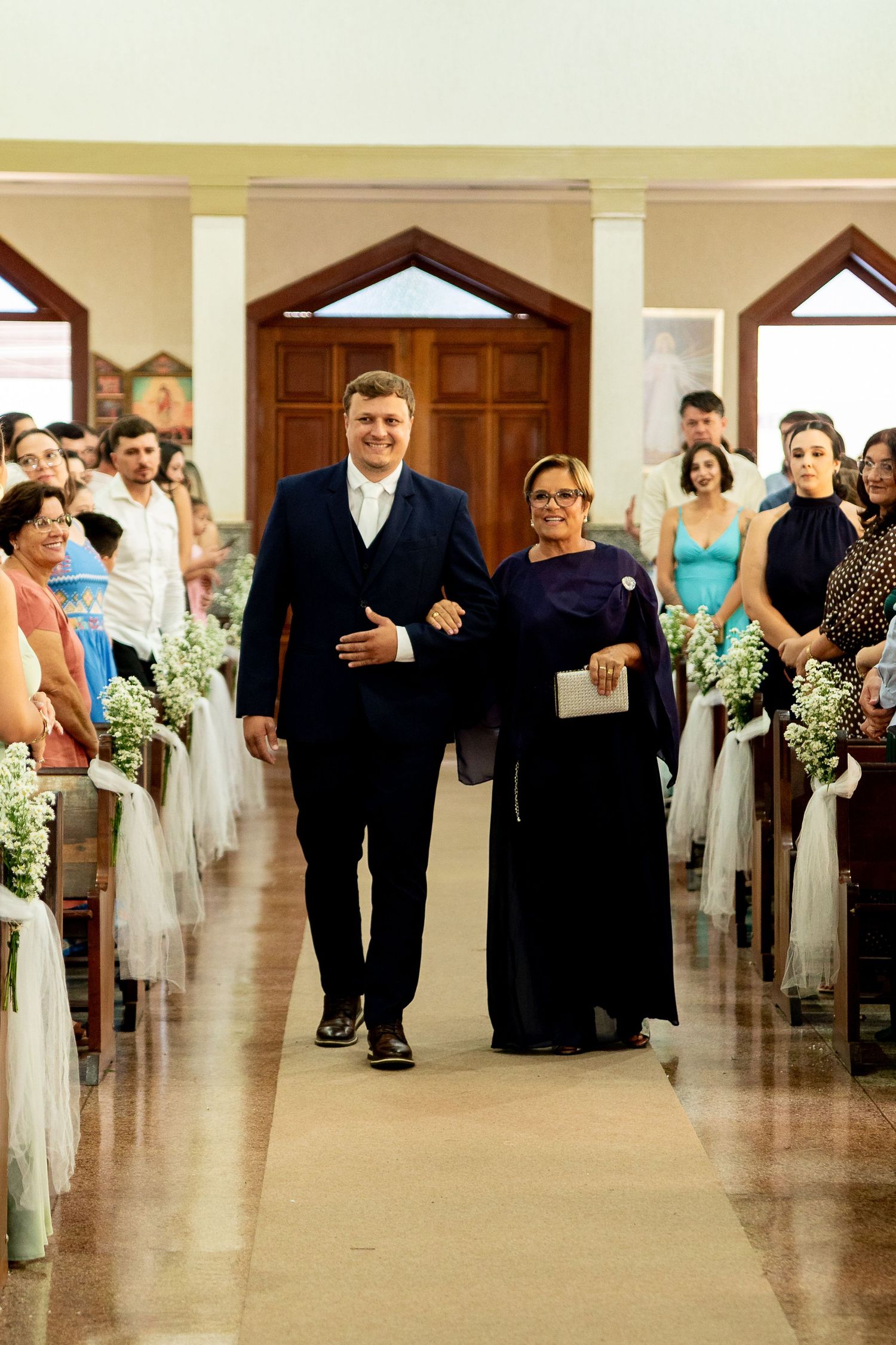 A groom in a navy suit walks down the church aisle with a woman in a purple dress during a wedding ceremony.