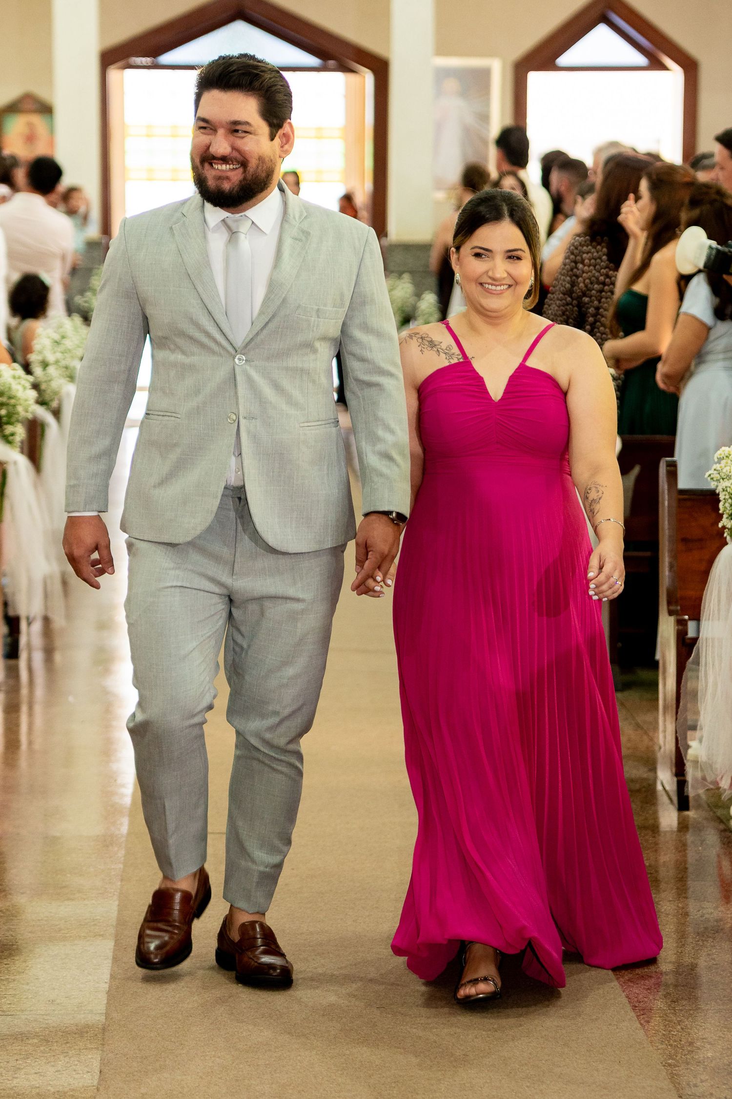 Couple walking down church aisle, man in gray suit, woman in pink pleated gown, holding hands at wedding.