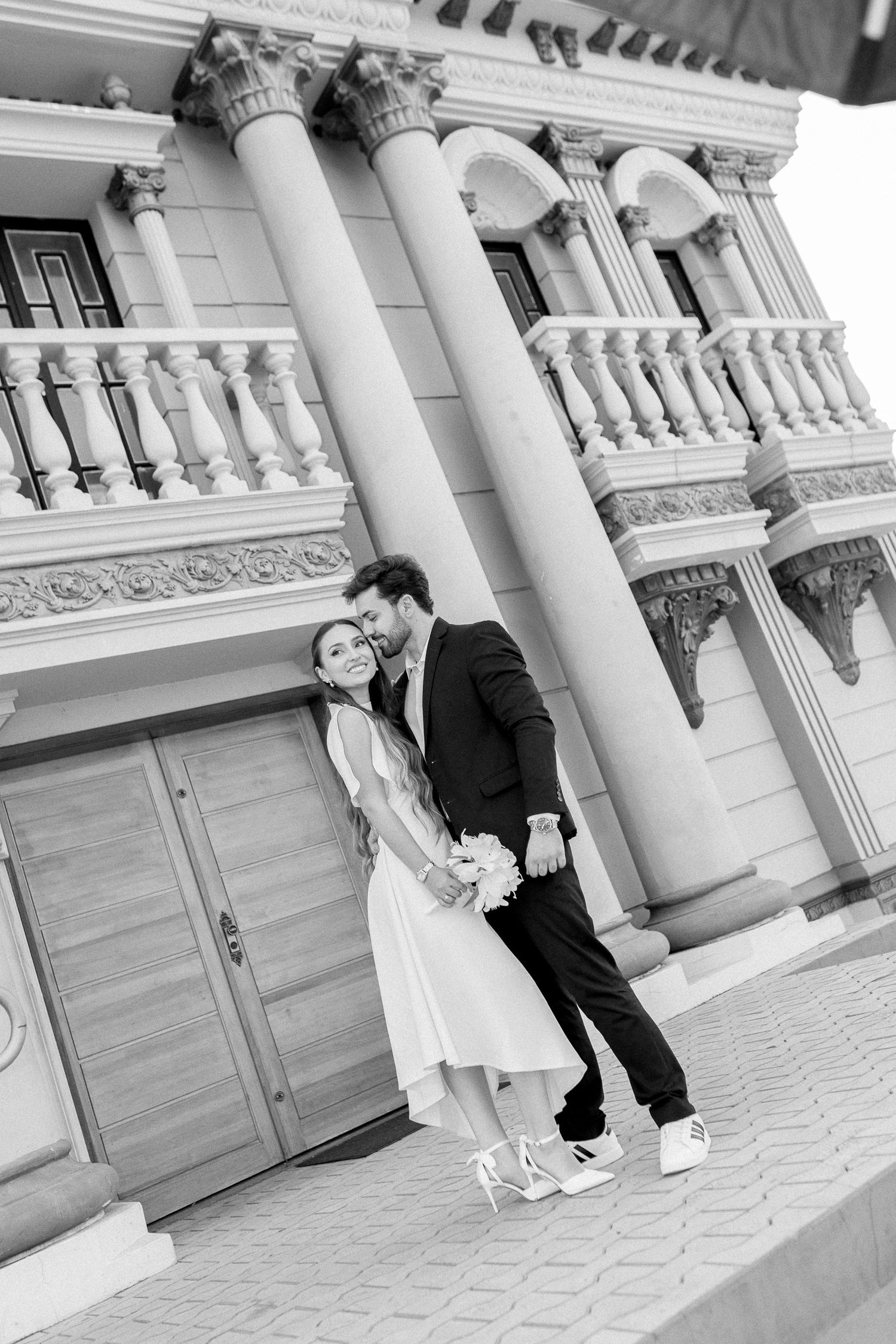 Black and white photo of a couple in wedding attire embracing before a grand classical building with ornate columns.