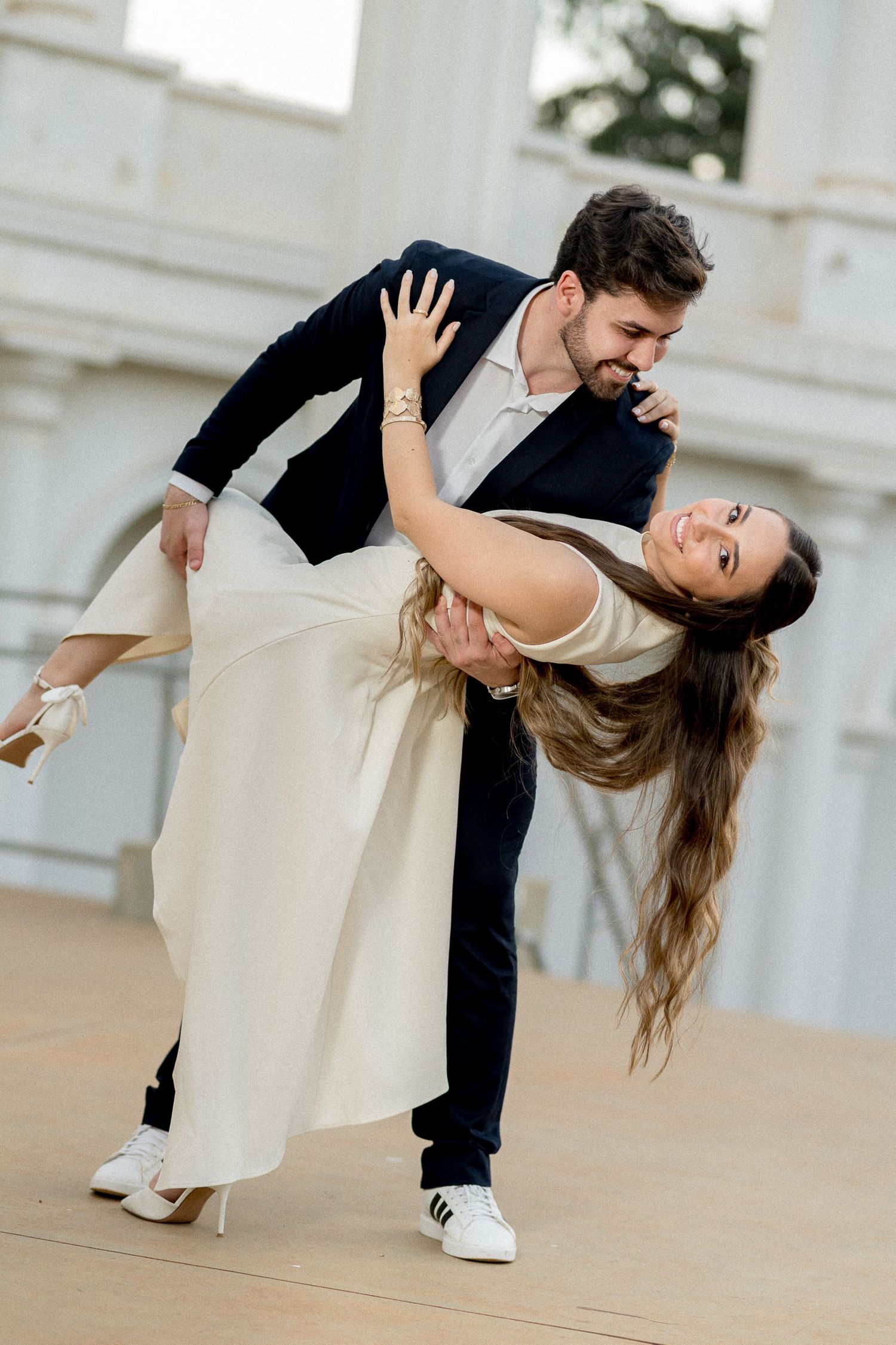 A couple elegantly dancing, man in navy suit dipping woman in white dress, at an outdoor venue.