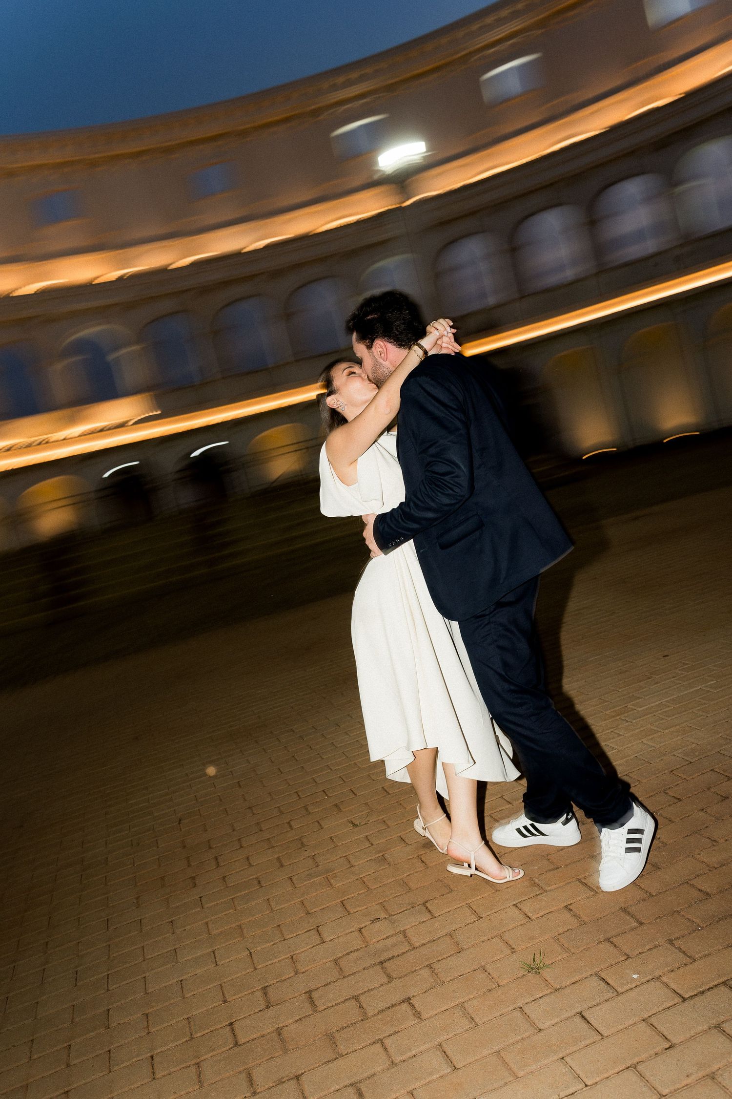 Couple sharing a romantic kiss at night near a historic colosseum-style building, woman in white dress, man in dark suit.