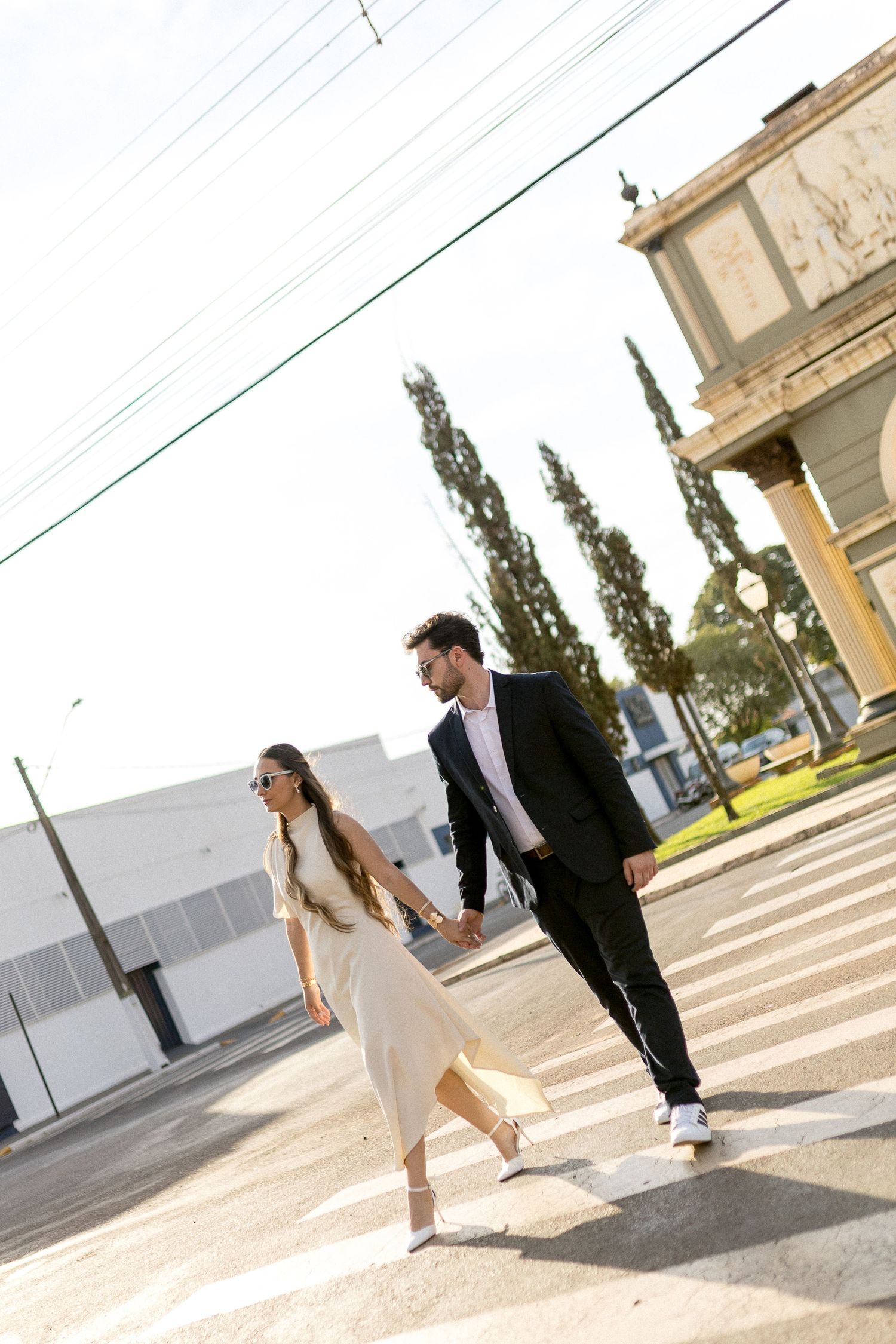 Stylish couple holding hands while crossing a zebra crossing on a sunny urban street near a classical building.
