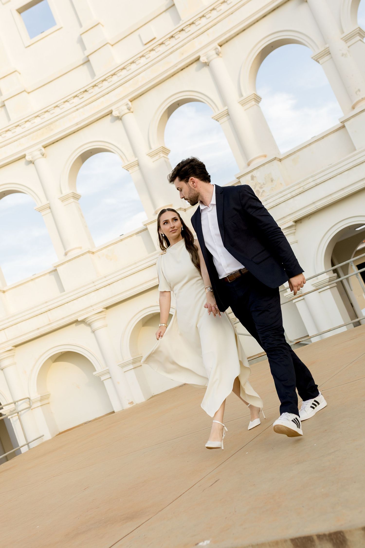 Couple walking hand in hand in front of the Colosseum in Rome, woman in white outfit, man in navy suit.