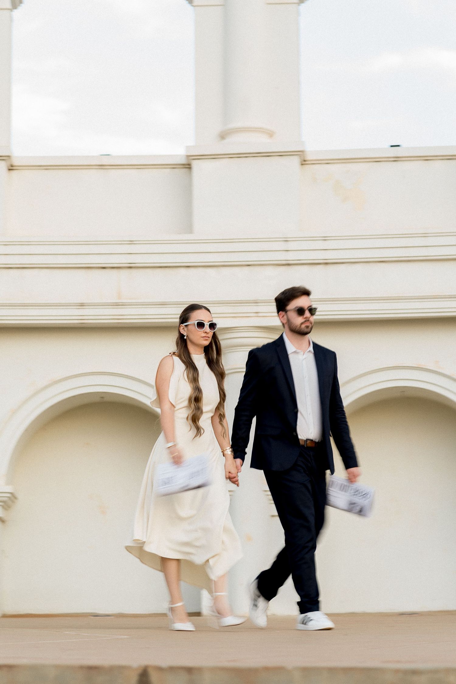 Stylish couple in sunglasses holding hands, woman in white dress, man in navy suit, walking near elegant white building.