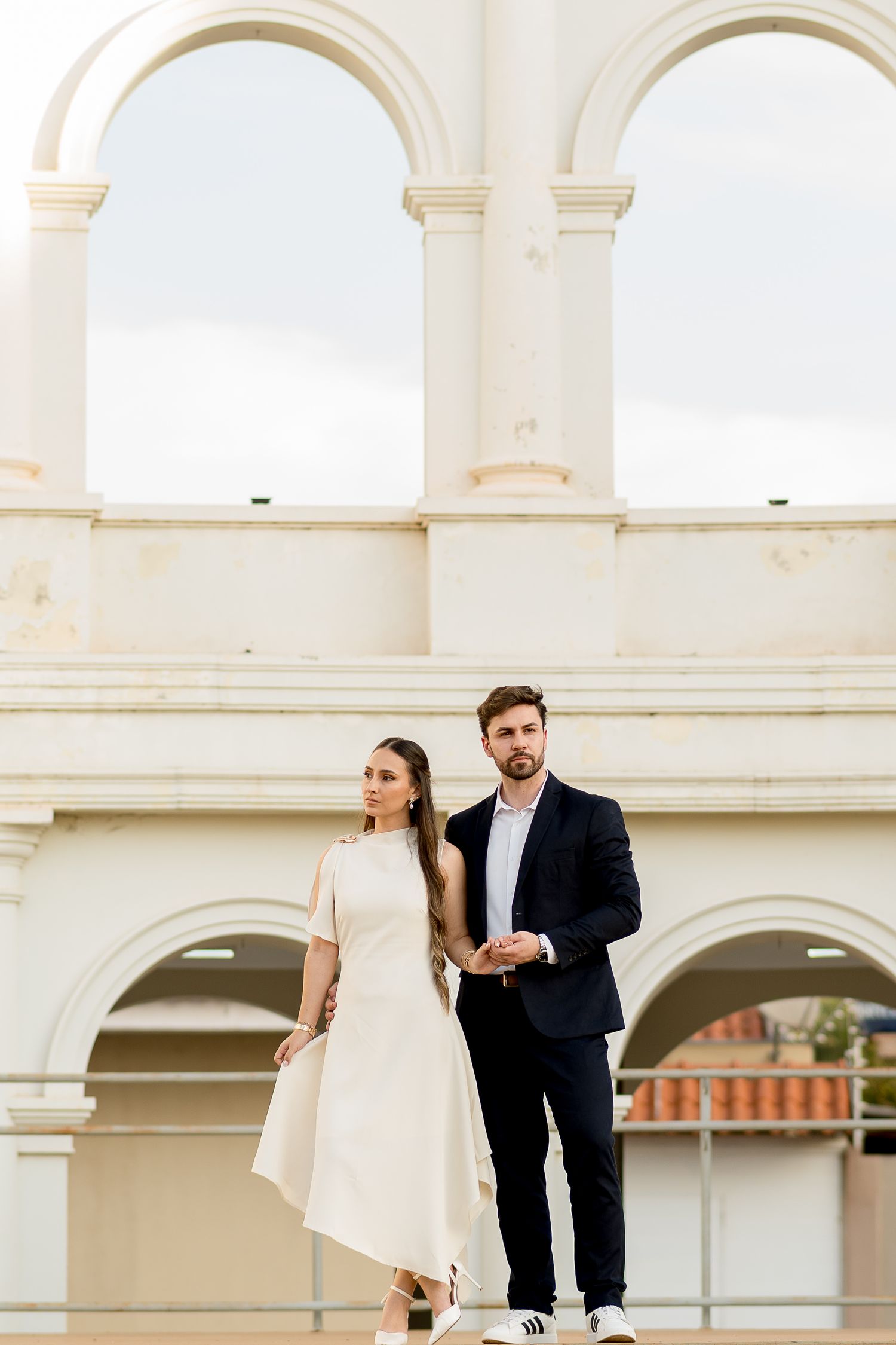 Elegant couple posing in front of white arched architecture, woman in cream dress and man in dark suit.