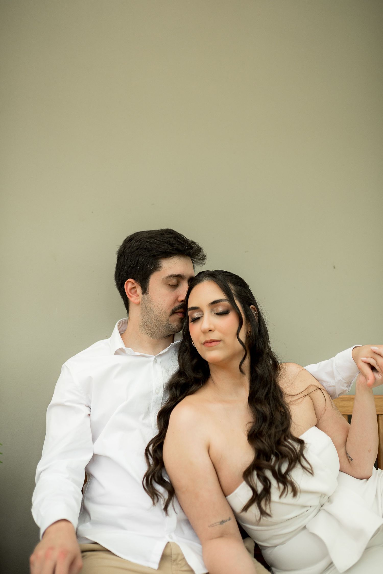 Romantic couple in white outfits sitting together, woman with long wavy hair resting against man's shoulder.