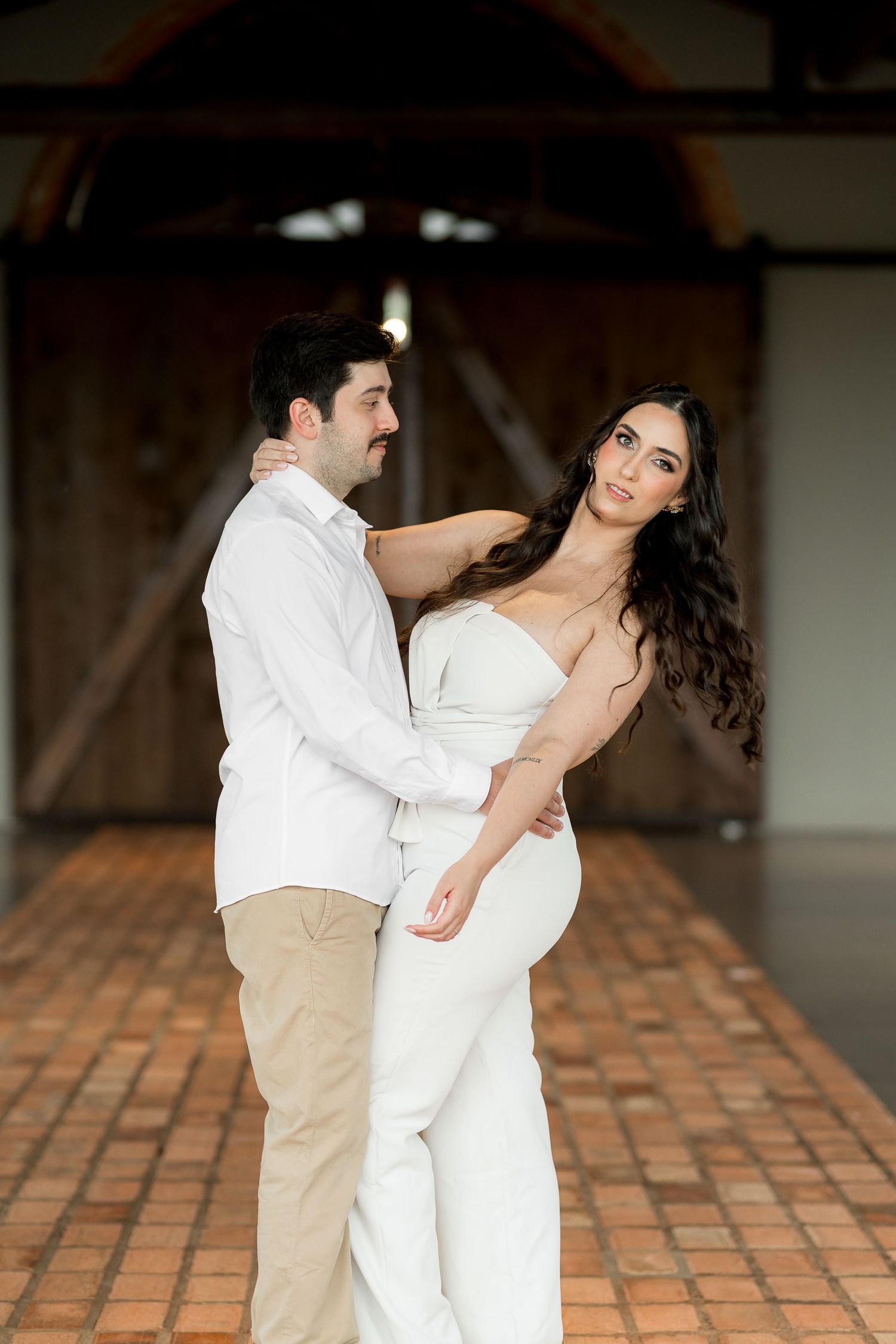 Couple in white attire embracing romantically in a rustic barn setting with brick floors and wooden doors.