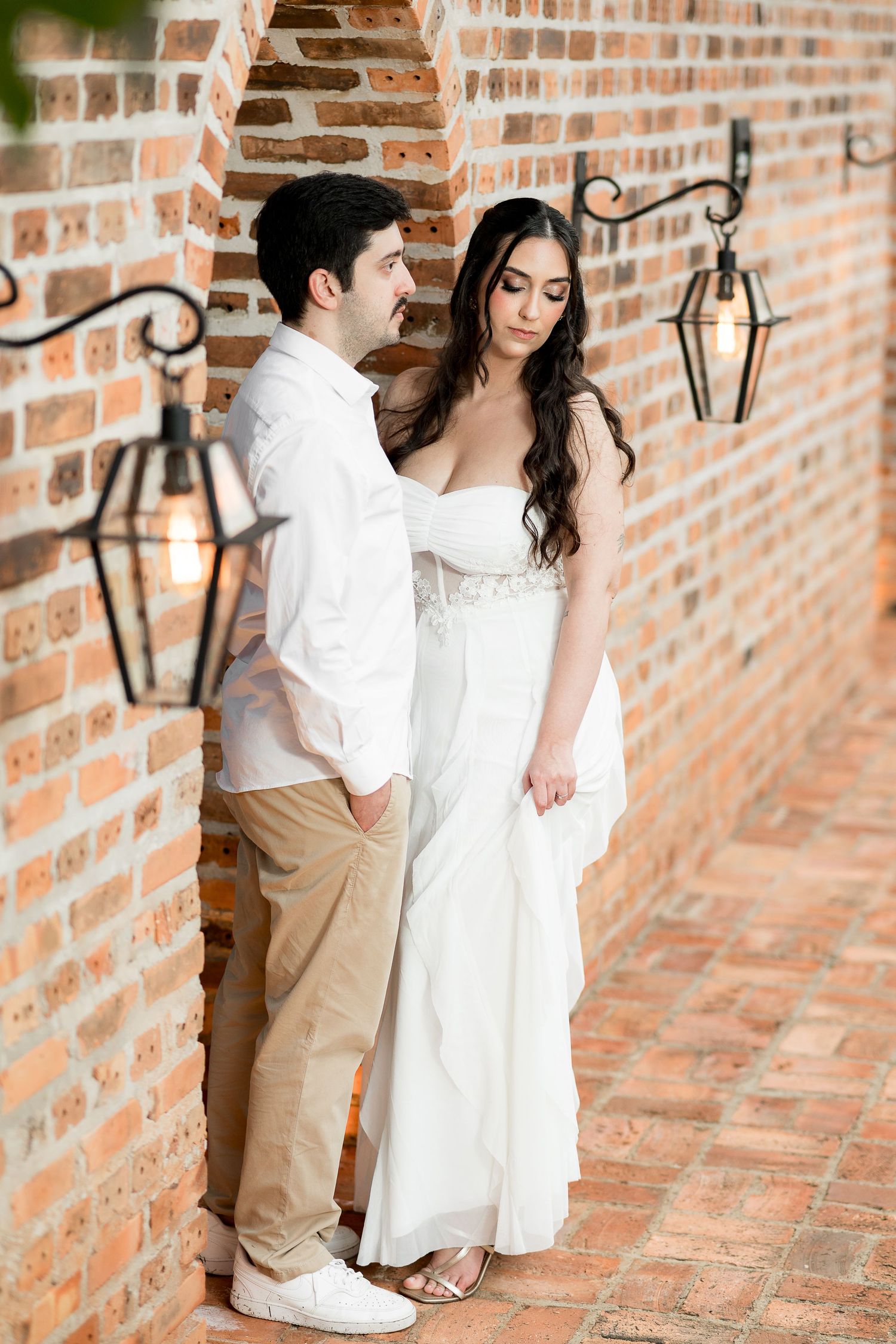 Elegant couple in wedding attire posing against rustic brick wall with vintage lanterns.