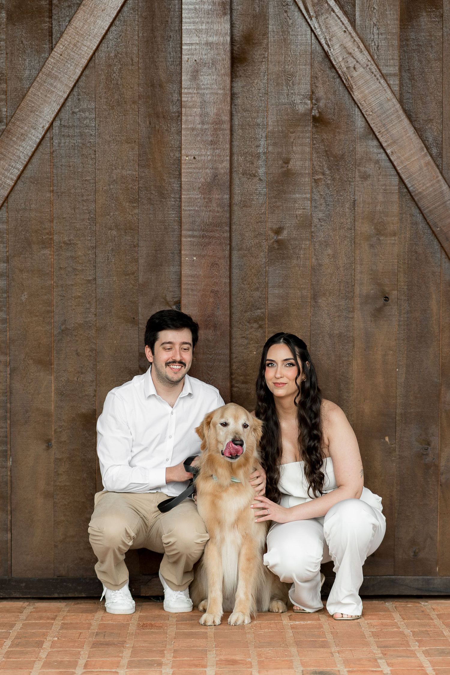 A smiling couple in white outfits crouches beside a golden retriever in front of rustic wooden barn doors.