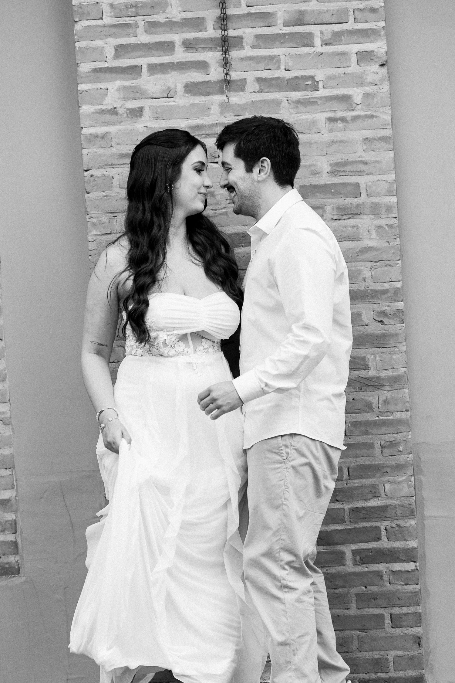 Black and white photo of a romantic couple in white attire sharing an intimate moment against a rustic brick wall.