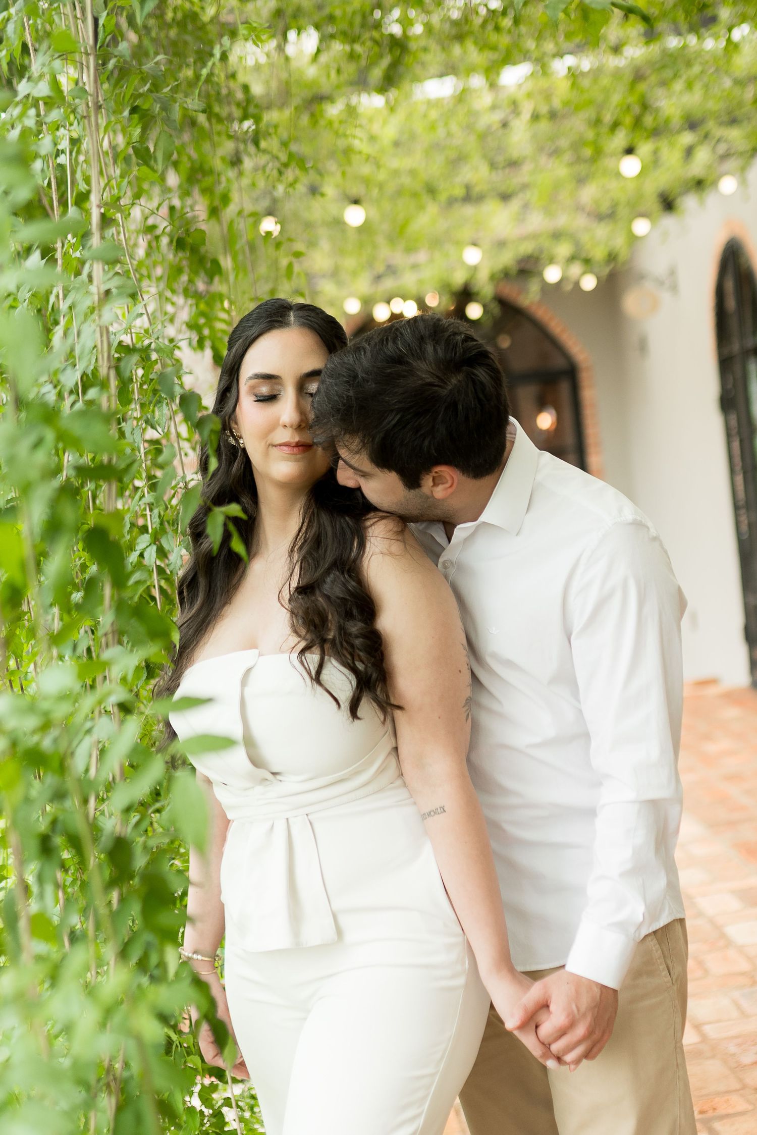 A couple in white attire shares a tender moment among lush green vines, the man gently kissing the woman's shoulder.