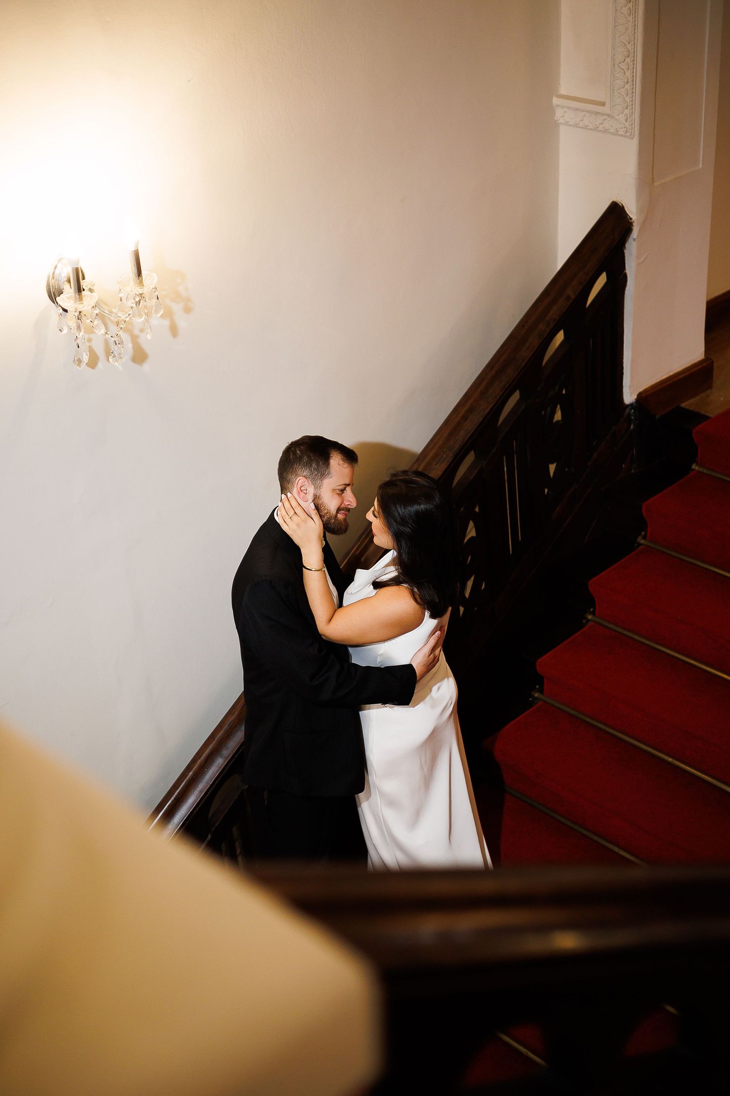 Bride and groom embracing tenderly on elegant red carpeted staircase with ornate wooden banister and wall sconce.