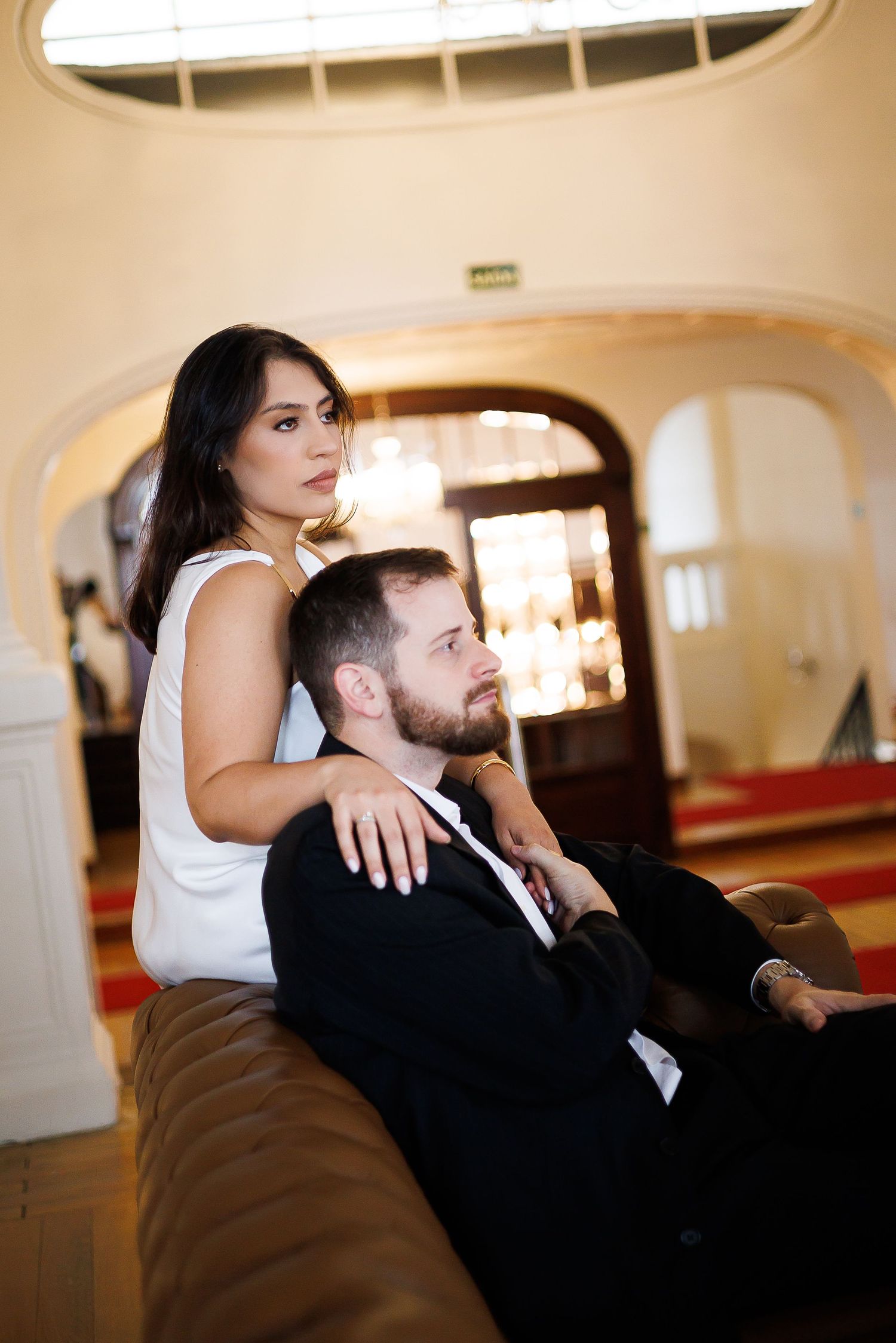 Elegant couple in formal wear posing in a luxurious interior, woman in white dress embracing seated man in black suit.