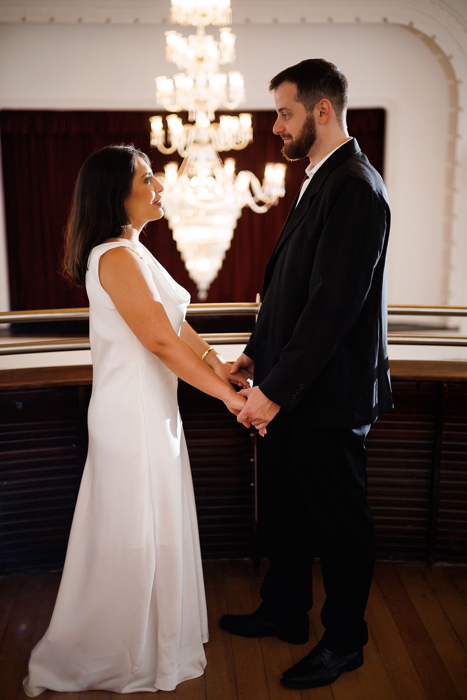 Bride and groom holding hands in elegant ballroom with chandelier, wearing white gown and black suit.