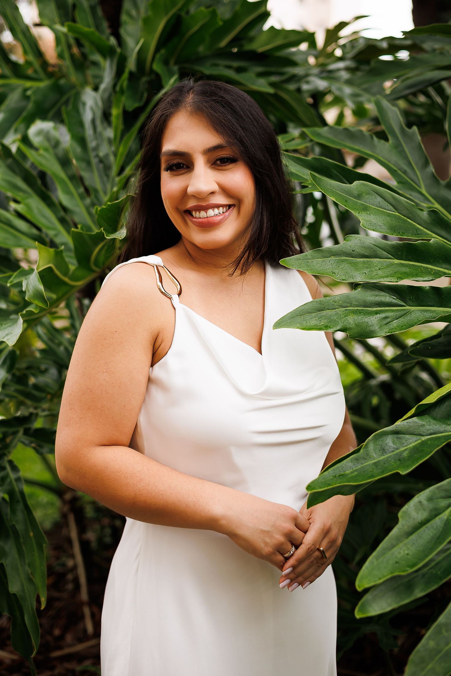 Smiling woman in white dress posing among tropical green plants outdoors.