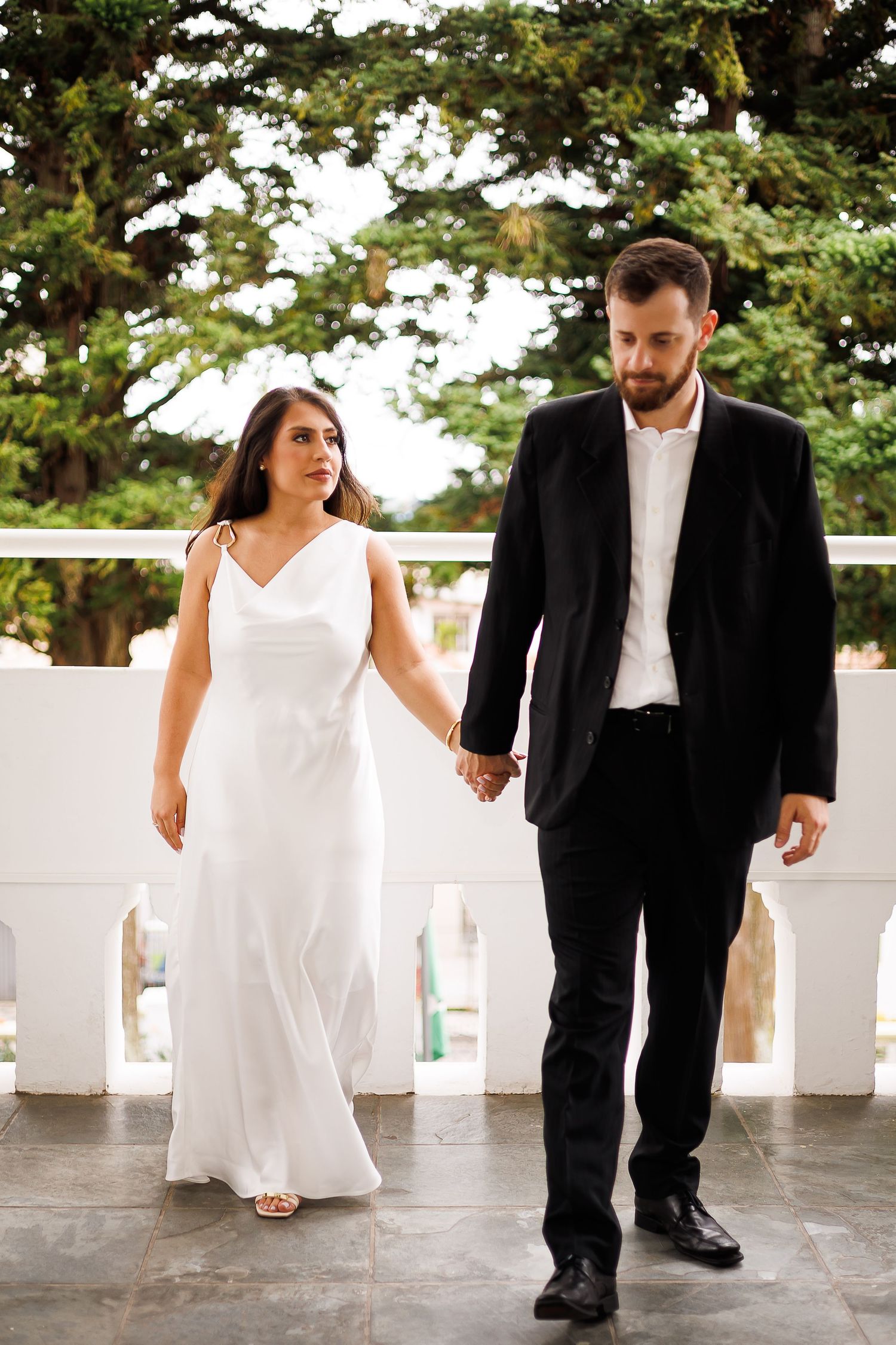 Elegant couple in formal attire holding hands on a terrace, bride in white gown and groom in black suit.