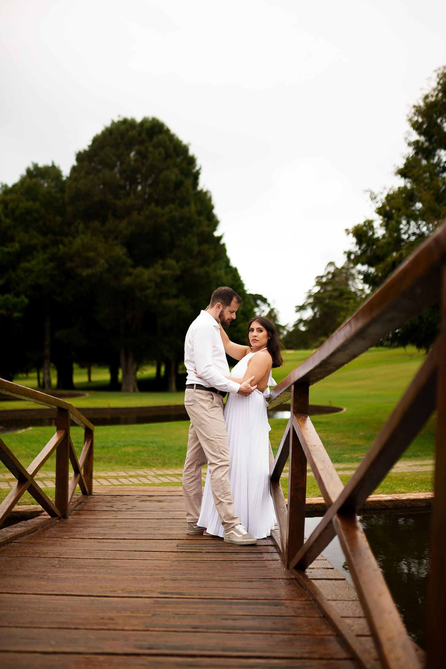 Couple in wedding attire embracing on a wooden bridge surrounded by lush green trees and grass.