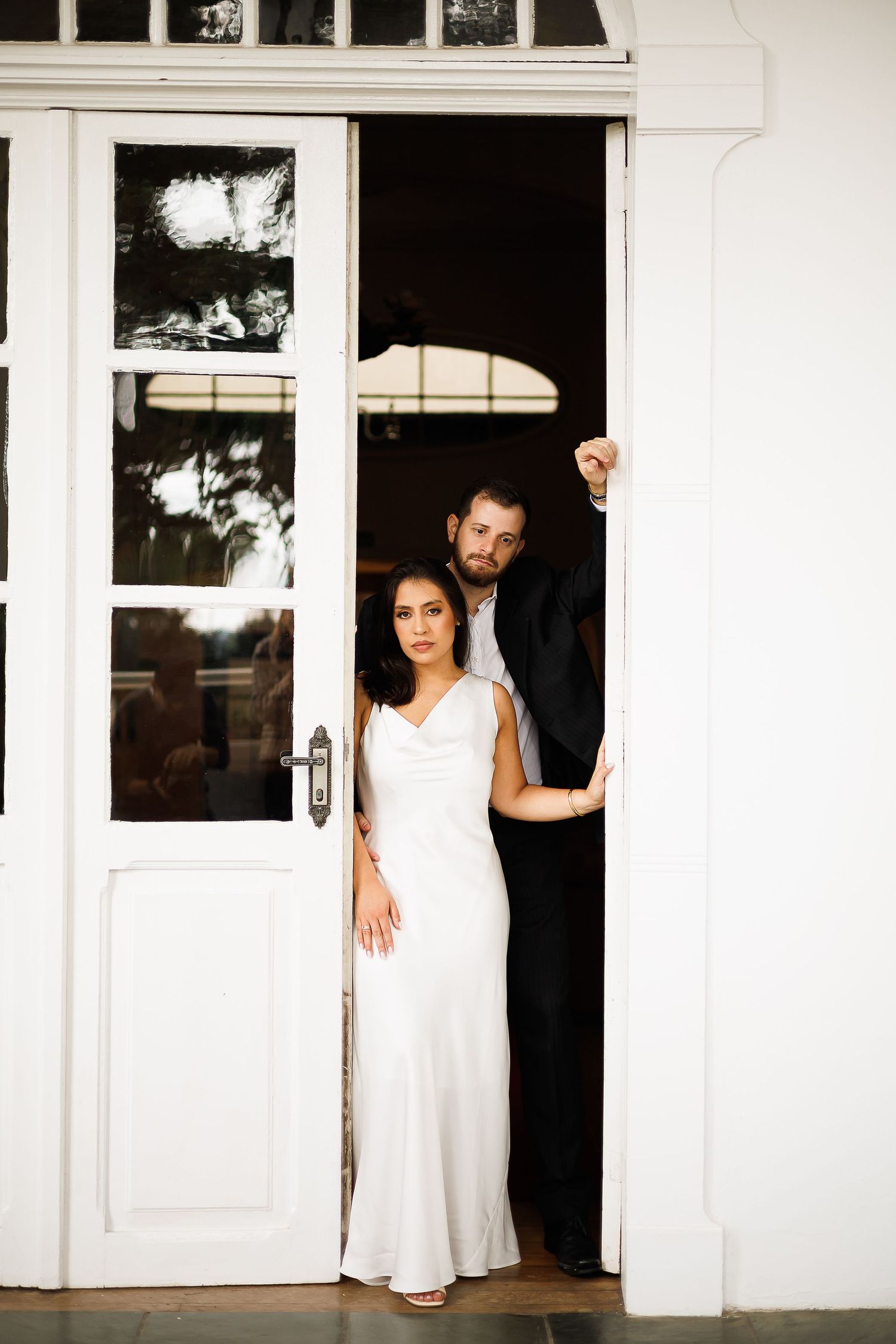 Bride in white gown and groom in black suit pose elegantly in white French doors at their wedding venue.