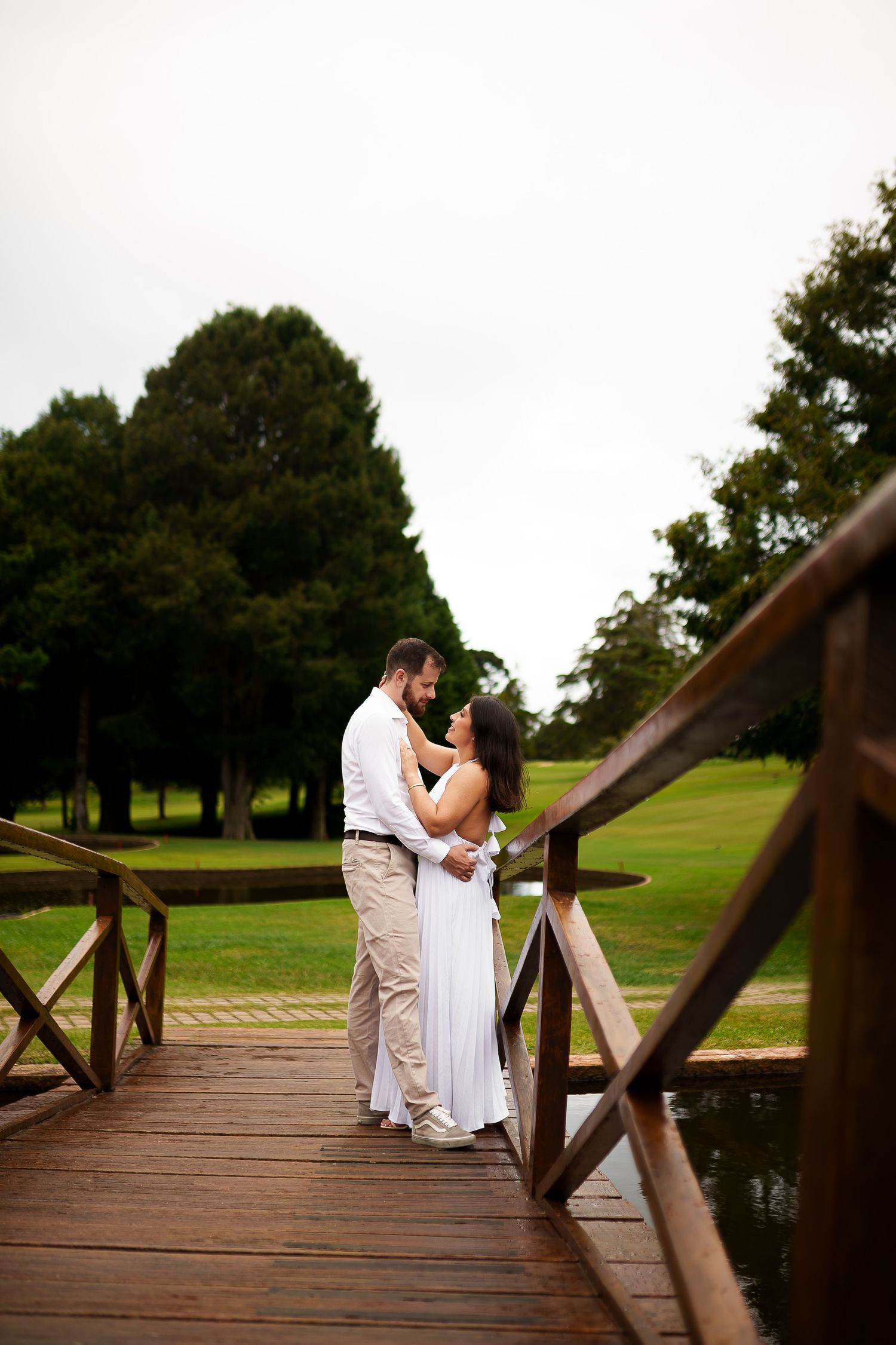 Couple in white wedding attire embracing on a wooden bridge surrounded by lush green trees and grass.
