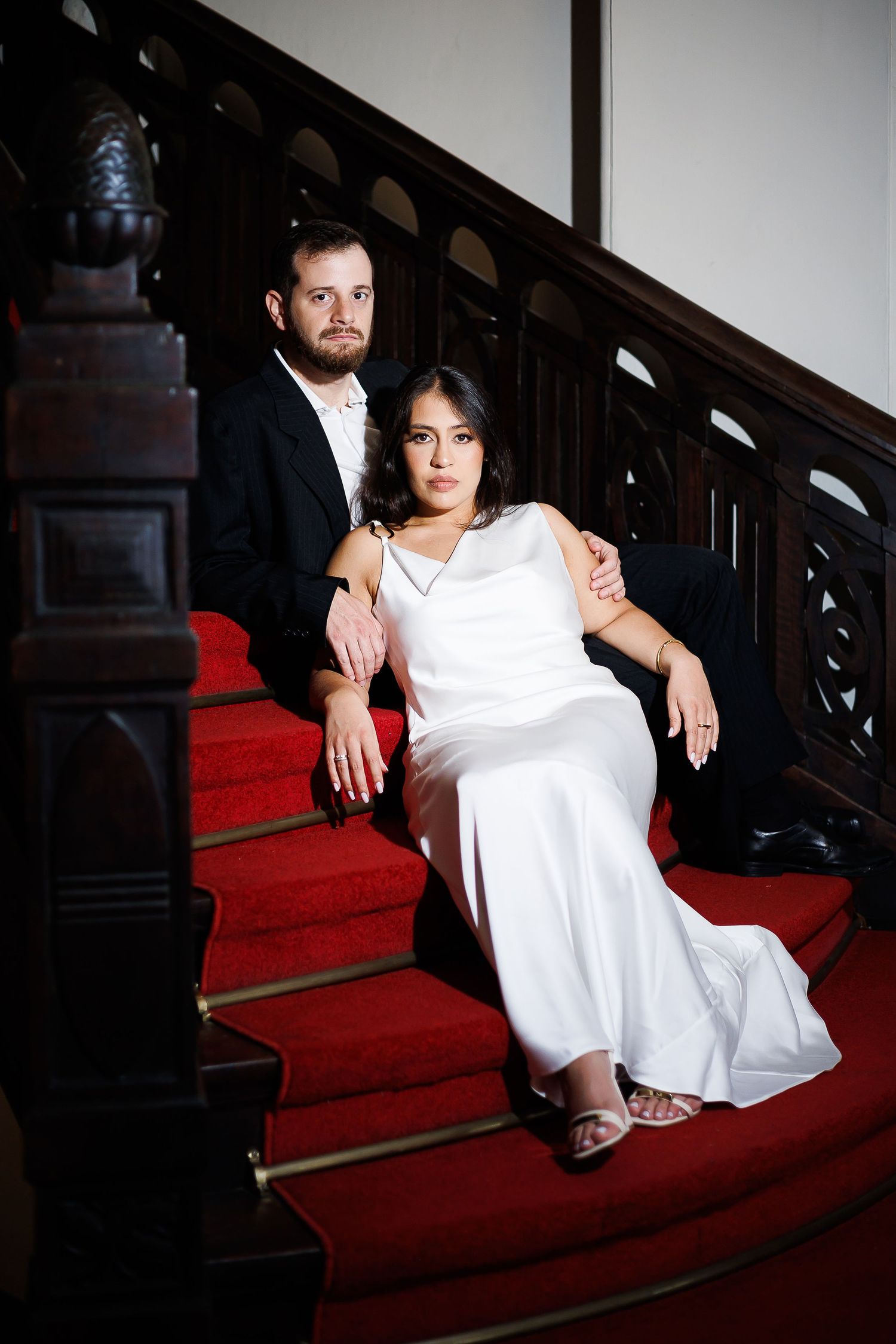 Bride in white gown and groom in black suit sit together on red-carpeted wooden staircase in elegant venue.