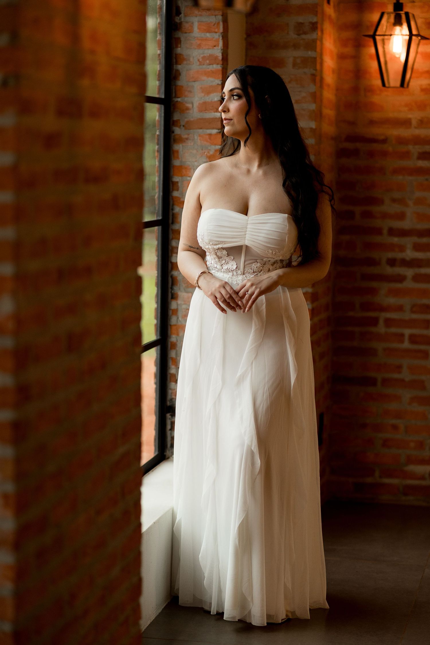 Elegant bride in a strapless white gown stands by a window in a rustic brick-walled venue.