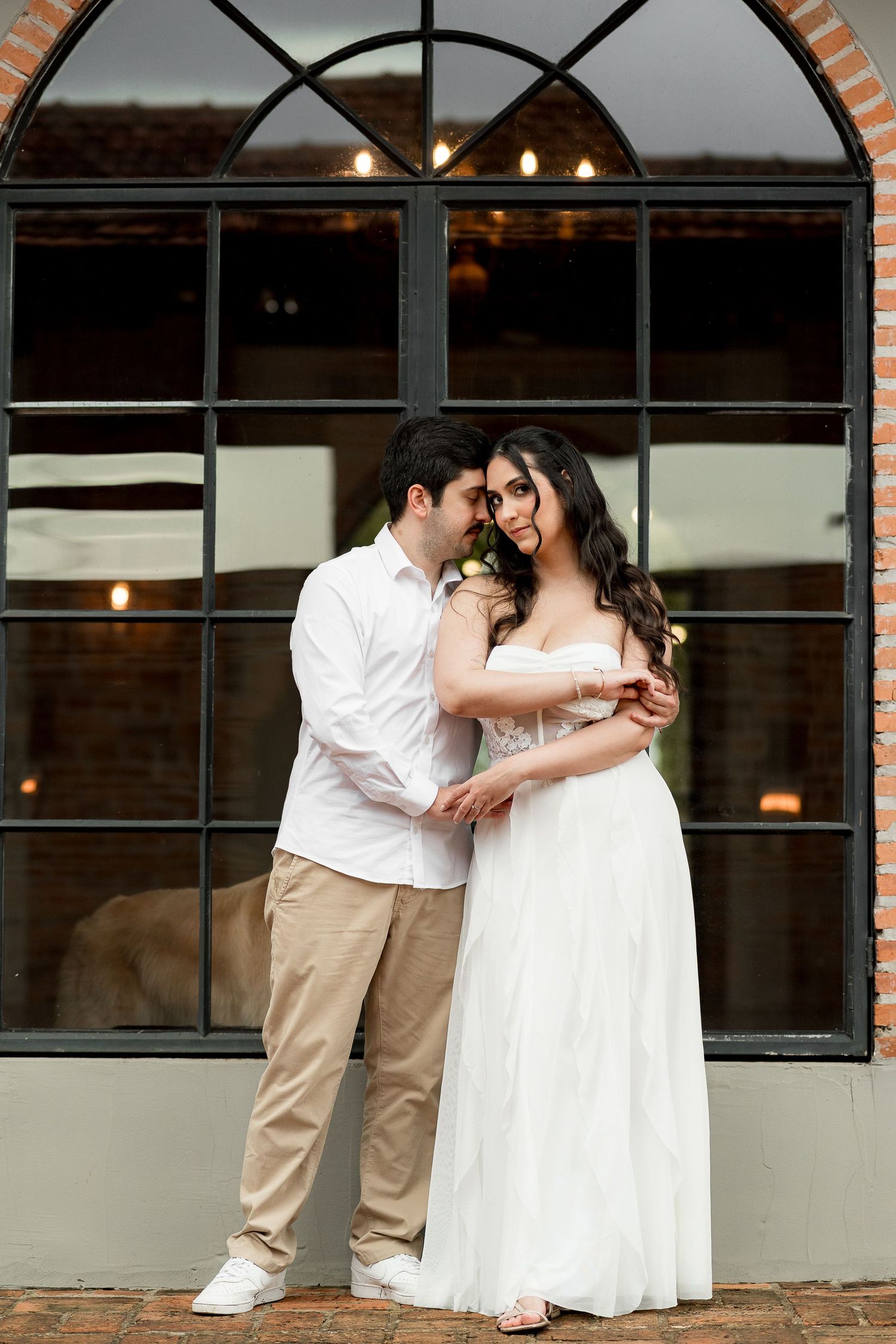 Couple in wedding attire embracing before large arched window, bride in white gown, groom in white shirt and khakis.