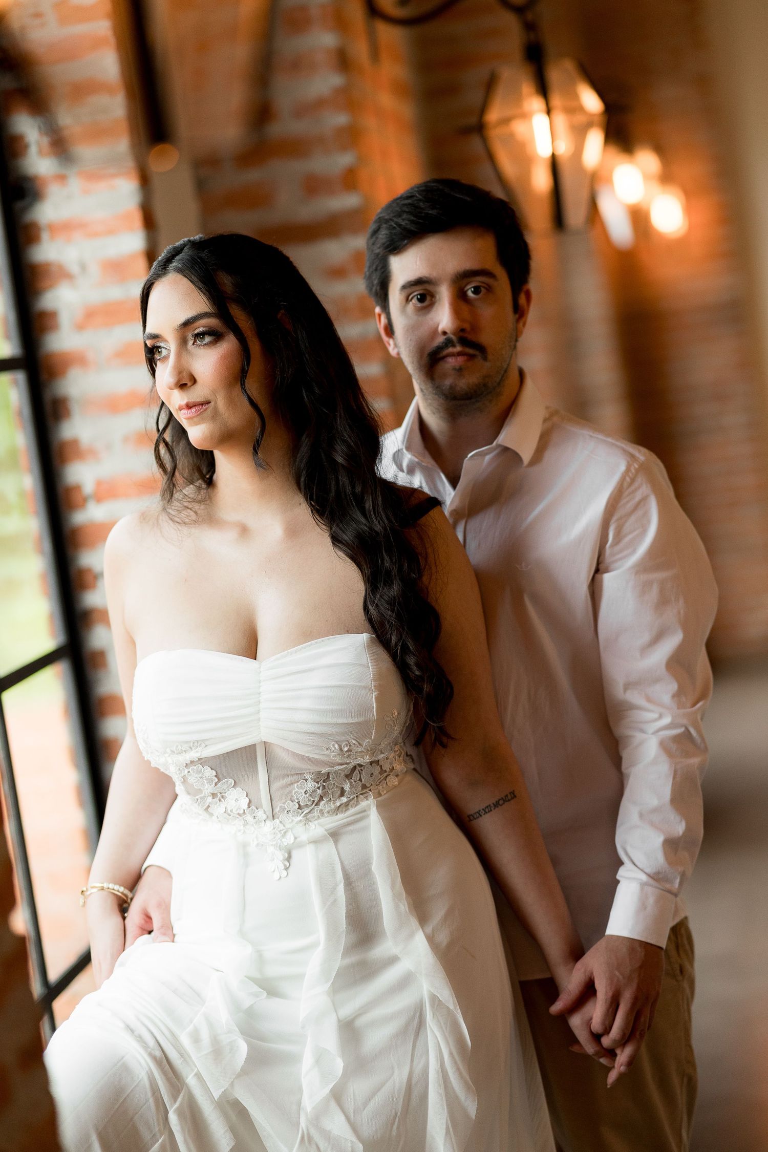 Elegant bride in strapless white gown holds hands with groom in white shirt against brick wall backdrop.