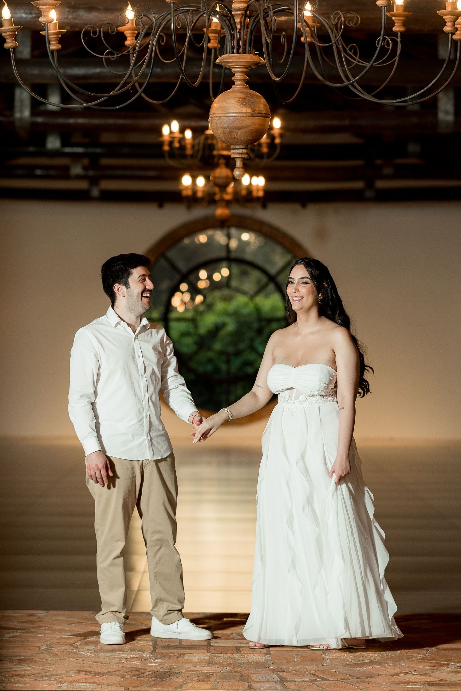 Couple holding hands and smiling under chandelier in elegant venue, woman in white gown, man in white shirt.
