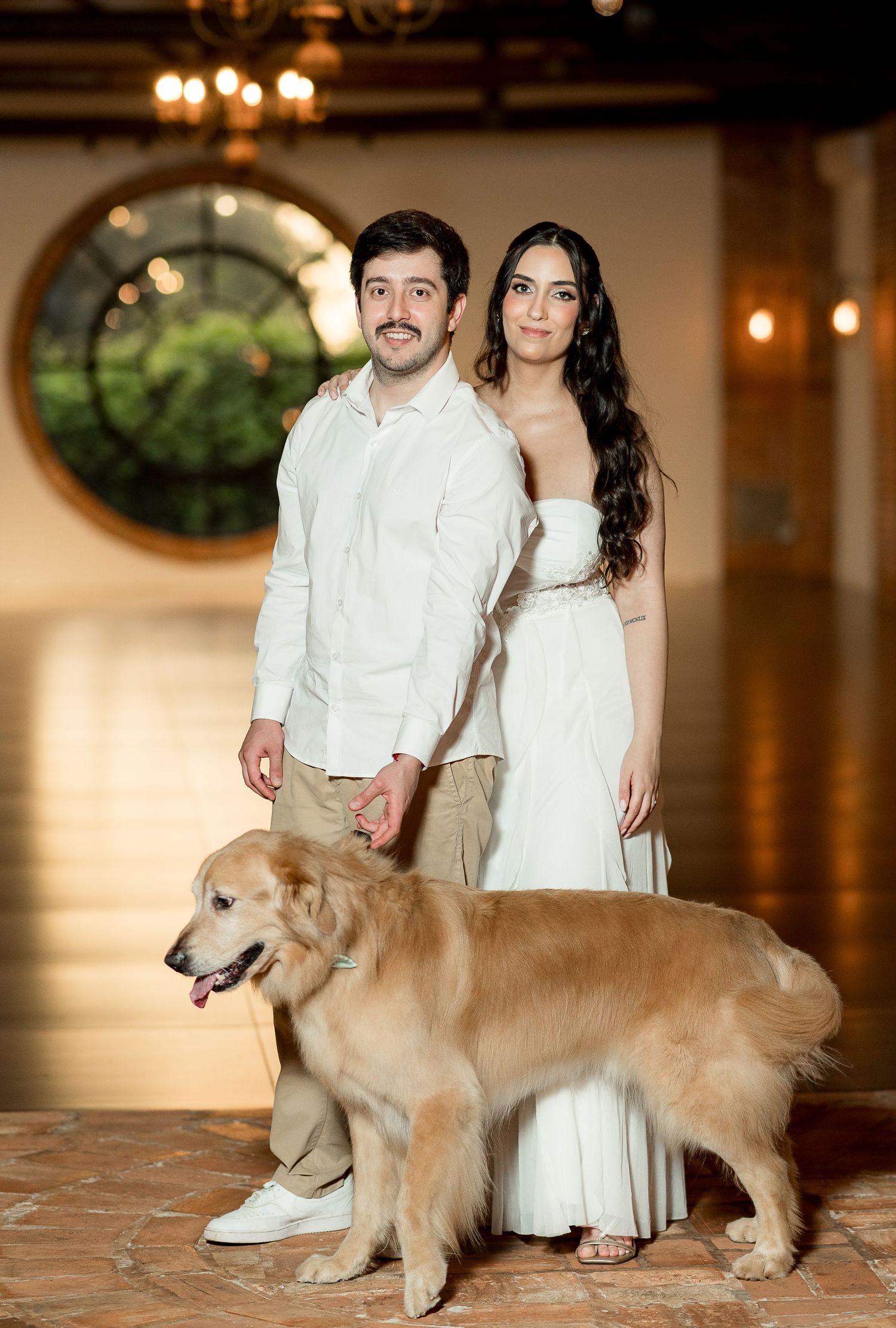 Couple in white wedding attire holding hands with a golden retriever dog in an elegant venue.