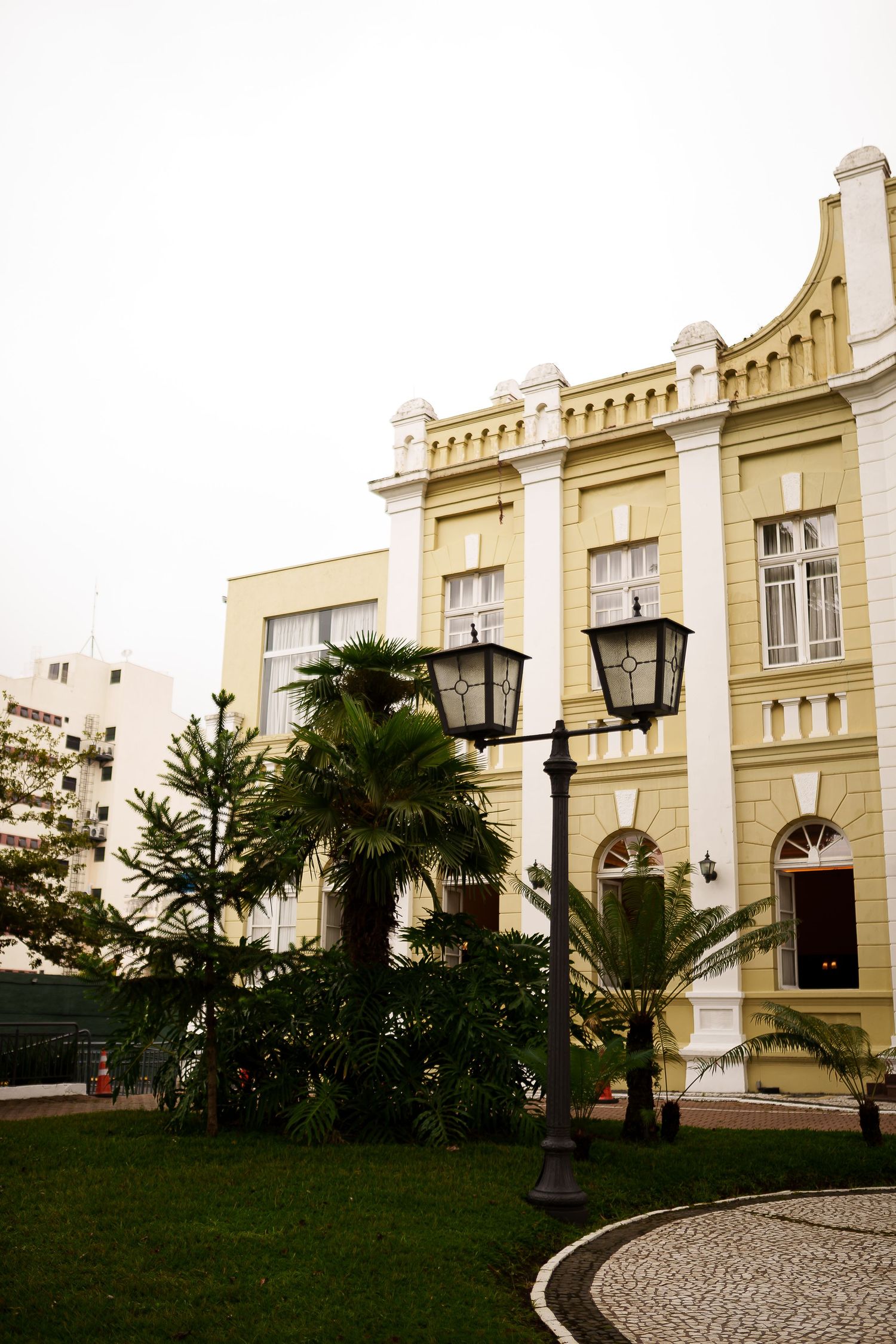 Yellow colonial building with ornate white trim, palm trees, vintage street lamp, and cobblestone path in a manicured garden.