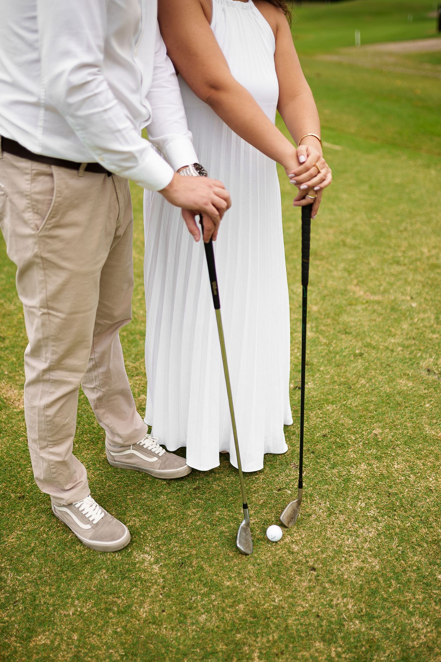 Couple holding golf clubs together on a green course, man in khaki pants and woman in white dress.