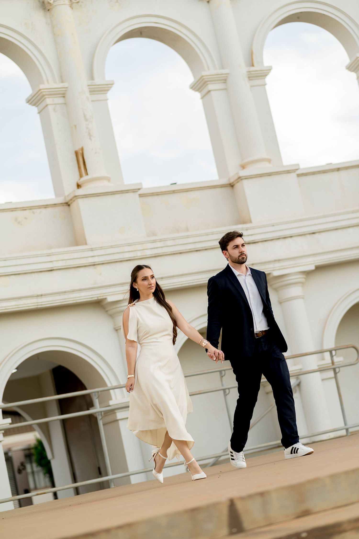 Elegant couple holding hands in front of white arched building, woman in cream dress, man in navy suit.