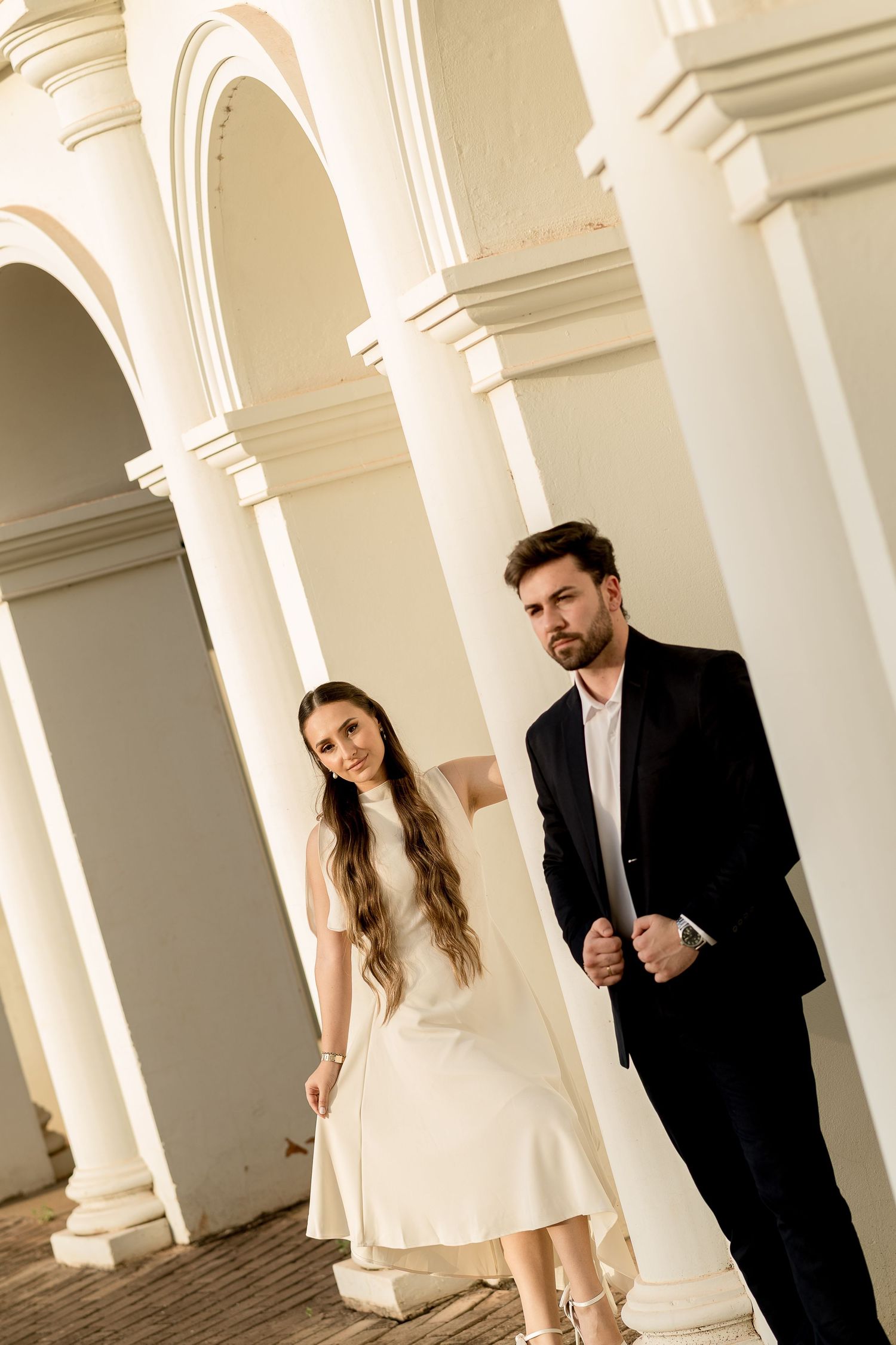 Elegant couple in formal attire posing between white classical columns and arches in a grand architectural setting.