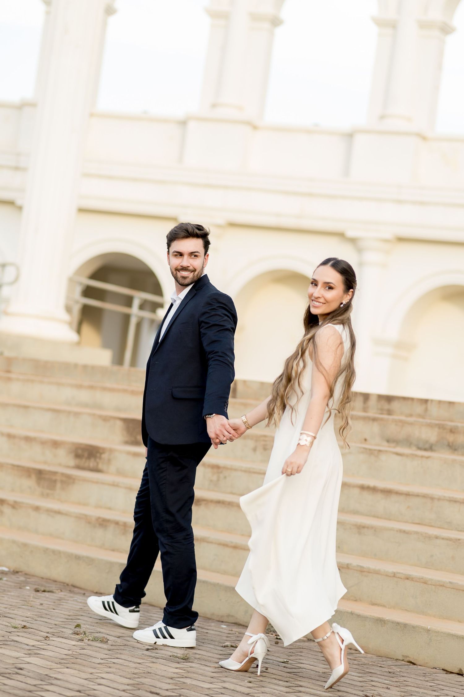 A smiling couple in formal attire holding hands on outdoor steps, woman in white dress, man in dark suit with sneakers.