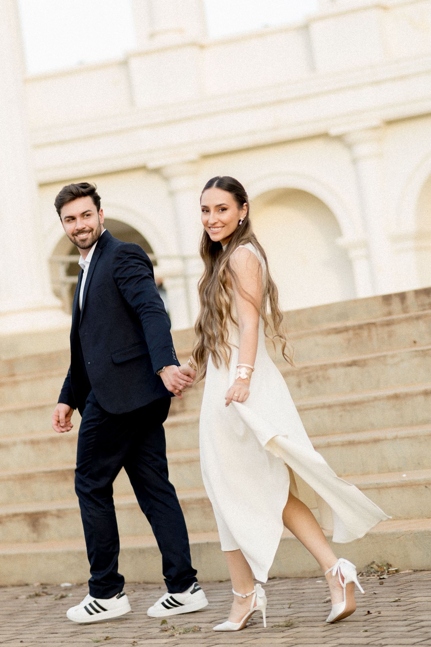 Smiling couple in elegant attire holding hands on stone steps, woman in white dress and man in navy suit.