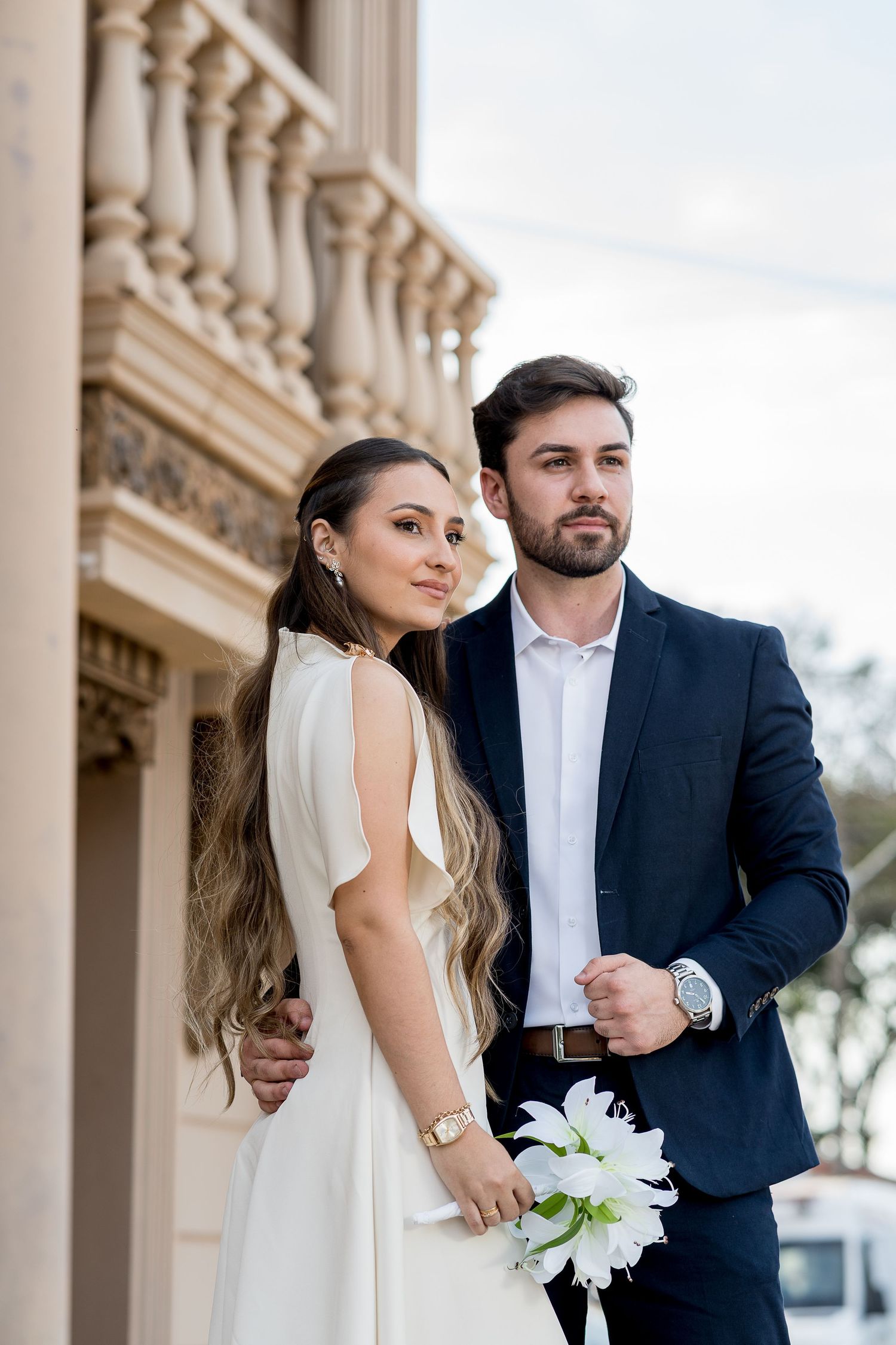 Elegant couple in wedding attire pose by classical columns, bride in white dress holding white flowers, groom in navy suit.