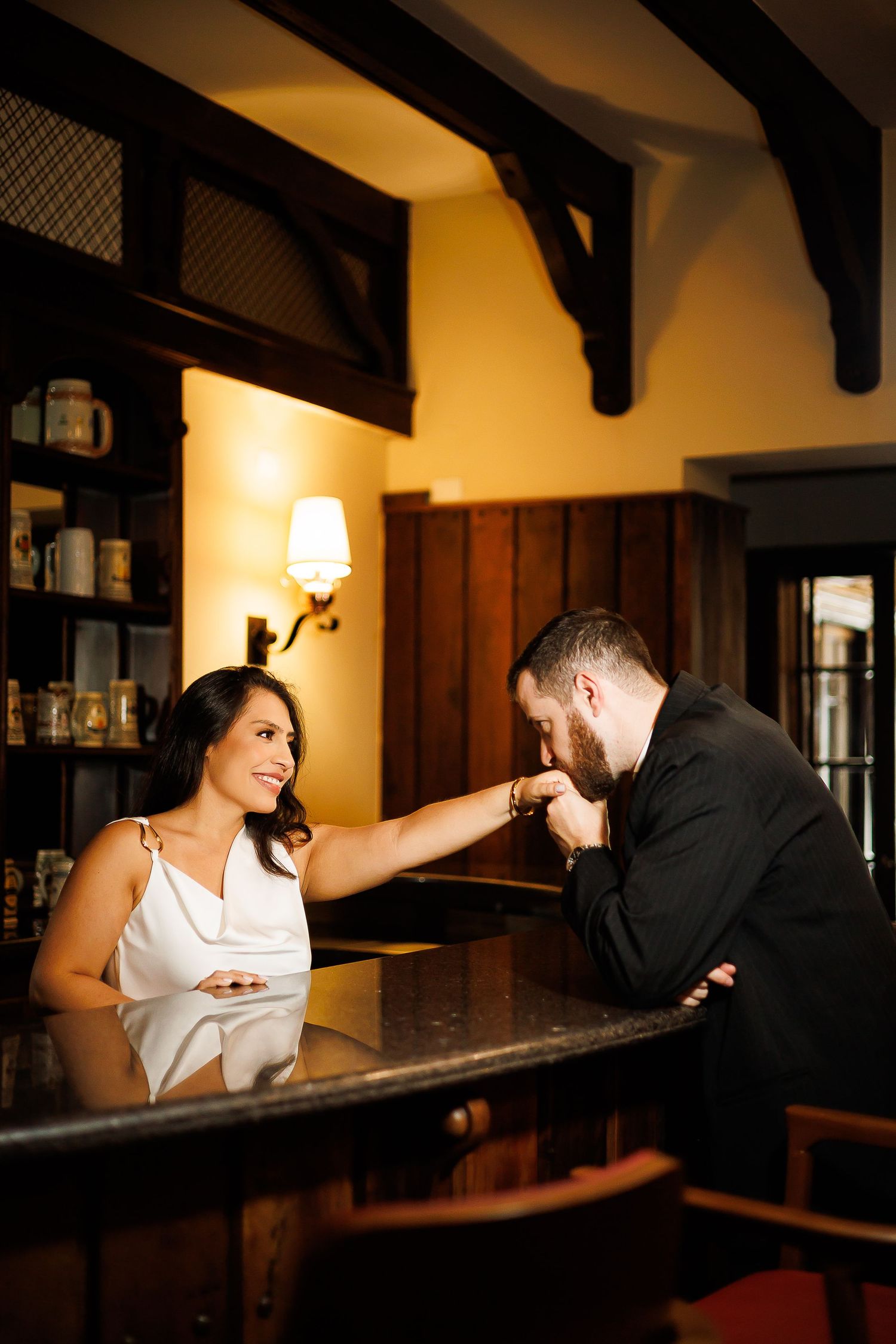 A man in a black suit kisses a woman's hand at an elegant bar, as she smiles warmly in a white dress.