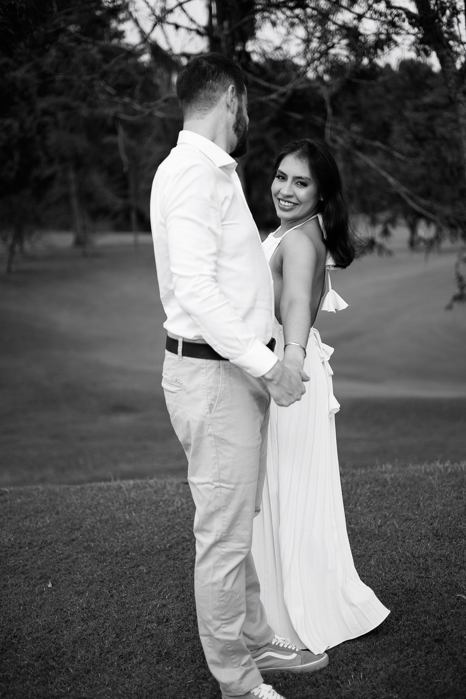 Black and white photo of a smiling woman in a white dress holding hands with a man in white outdoors.