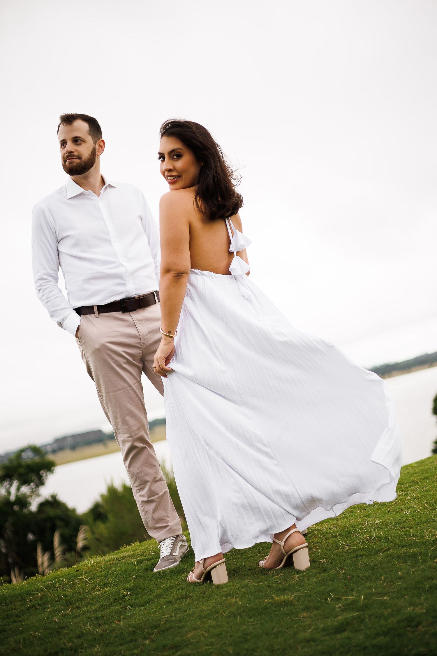 Couple in white outfits standing on green grass near water, woman in flowing white dress smiling over shoulder.