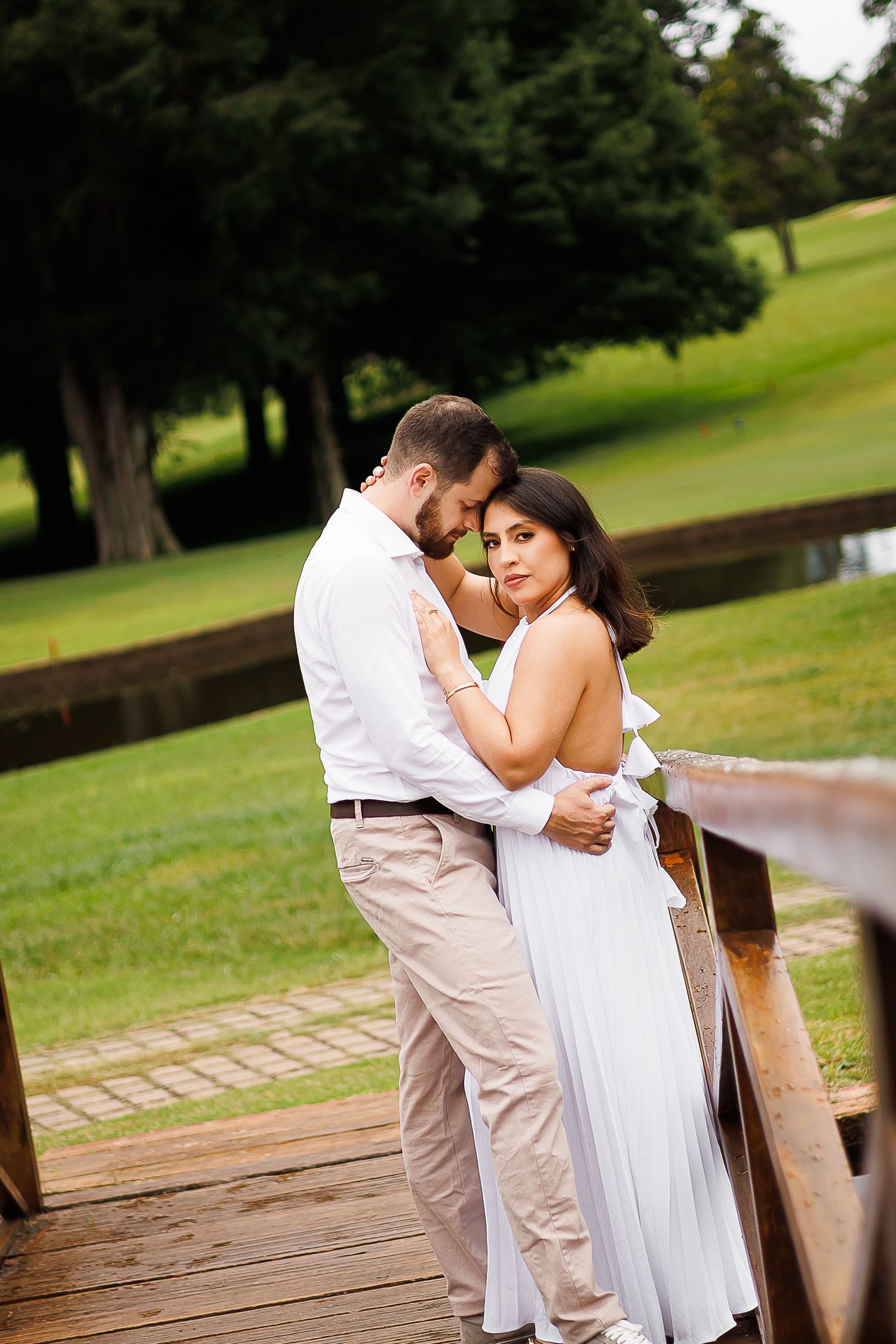 Romantic couple embracing on a wooden bridge in a lush green park, woman in white dress, man in white shirt.