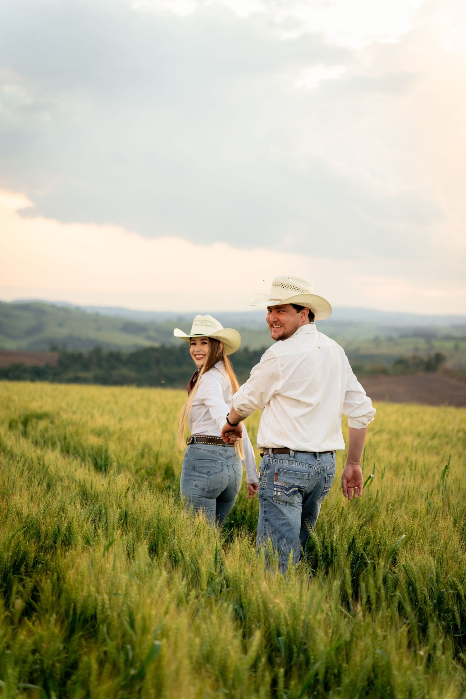 A smiling couple in cowboy hats and jeans hold hands while walking through a lush green wheat field at sunset.