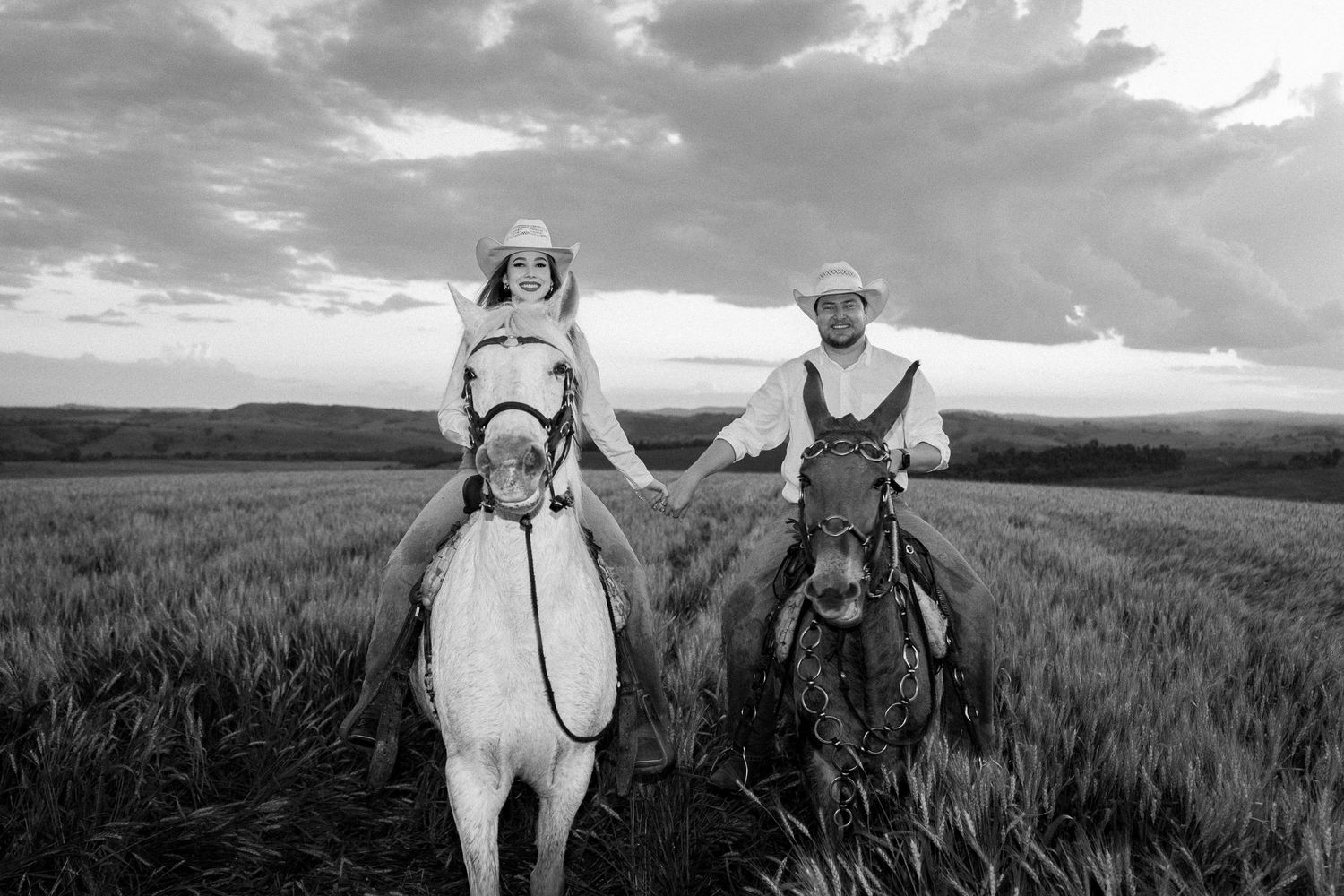 Black and white photo of a couple in cowboy hats riding horses through a wheat field under dramatic cloudy skies.