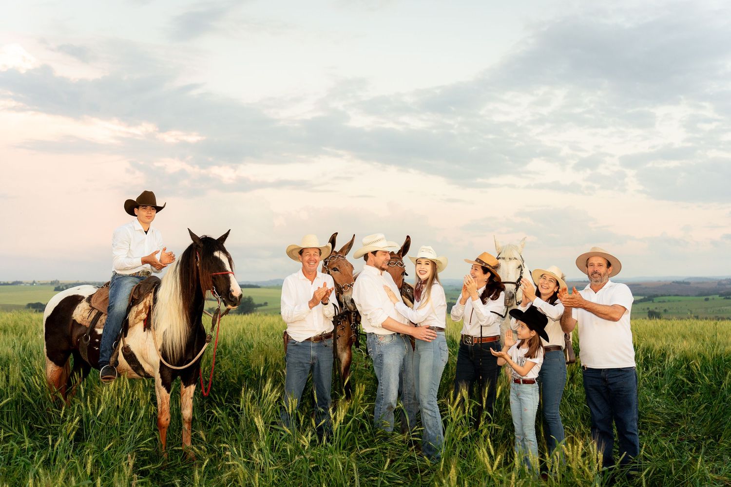 A group of people in cowboy hats pose in a green field, one riding a horse, under a pastel sunset sky.