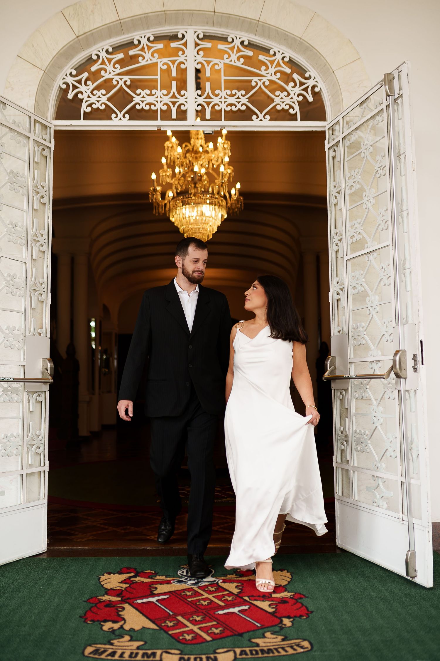 Bride and groom exit ornate venue doors under golden chandelier, walking on crest carpet.