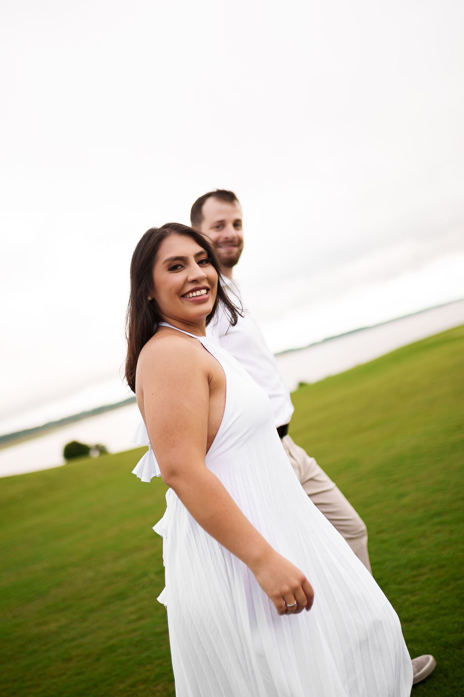A smiling couple in white attire walks on a green lawn near water, the woman turning back joyfully.